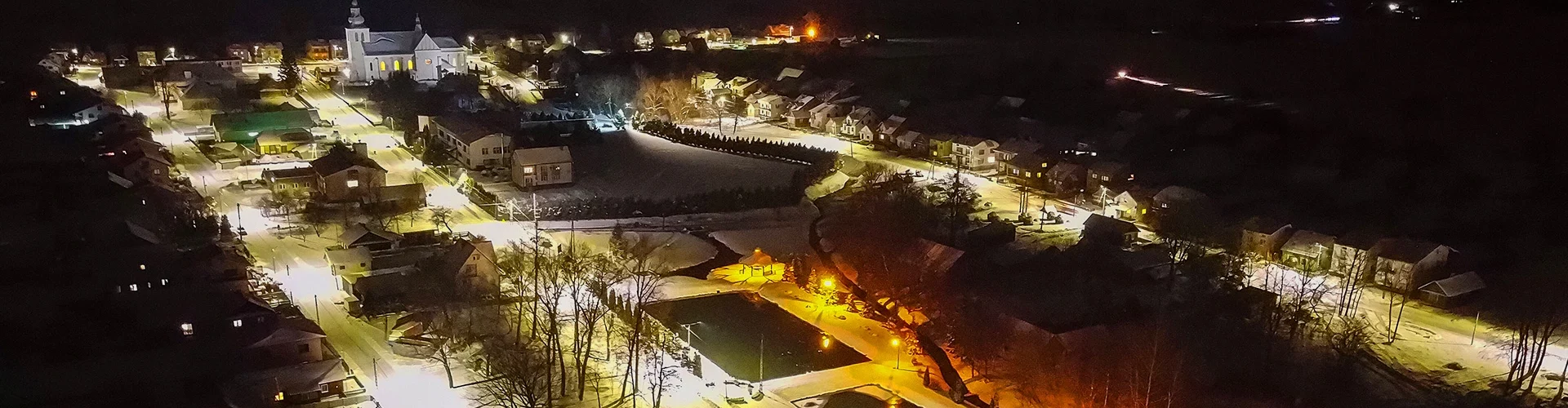 Nocny widok na oświetlone ulice miasteczka z białym kościołem, domami i drzewami na tle ciemnego nieba.