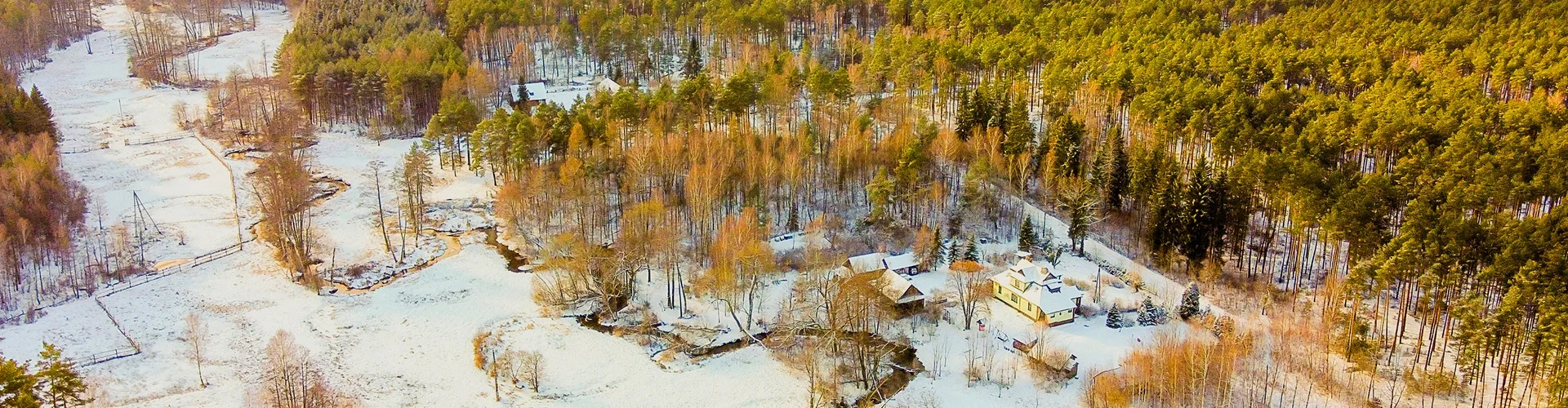 Zima w lesie z domem i budynkami pokrytymi śniegiem, wokół drzewa iglaste i liściaste, częściowo ośnieżona polana, strumień.