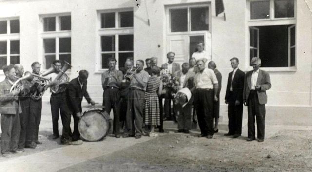Grupa mężczyzn i kobieta z bukietem kwiatów stoją przed jasnym budynkiem z trzema oknami i drzwiami, obok nich mężczyzna uderza w duży bęben, kilku gra na instrumentach dętych.