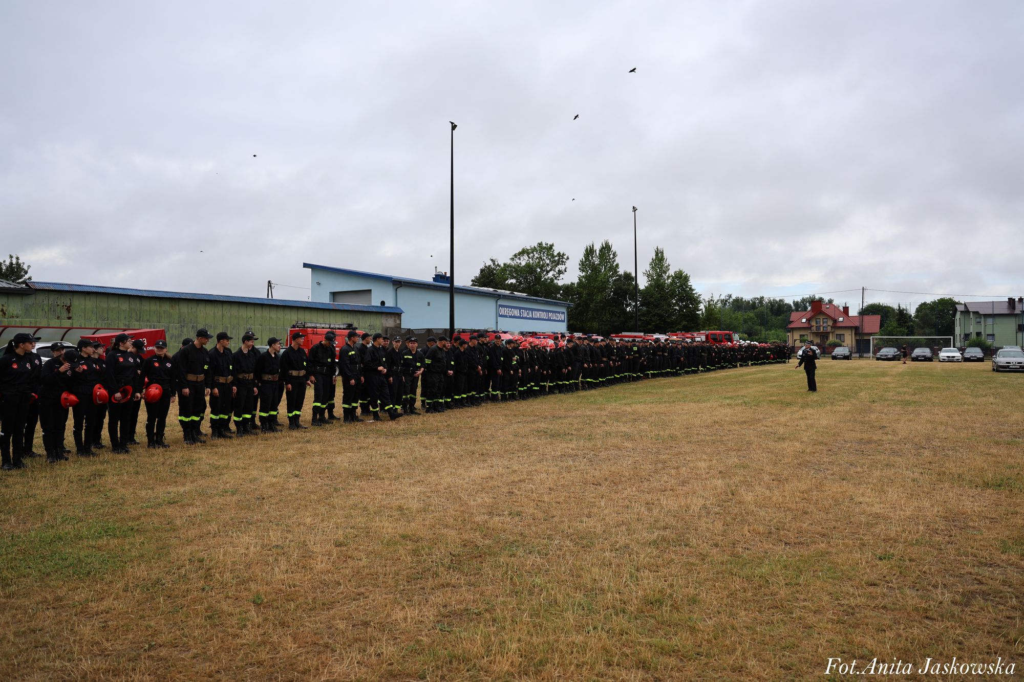 Duża grupa osób ubranych w czarne mundury i czerwone hełmy stoi w szeregu na trawie przed budynkiem i czerwonymi pojazdami ratowniczymi pod pochmurnym niebem.