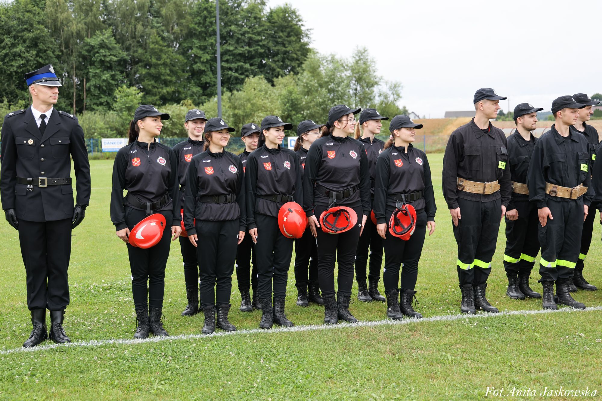Grupa osób w mundurach stojących na zielonej trawie, większość w czarnych strojach z czerwonymi hełmami w rękach, jedna osoba w granatowym mundurze z czapką.
