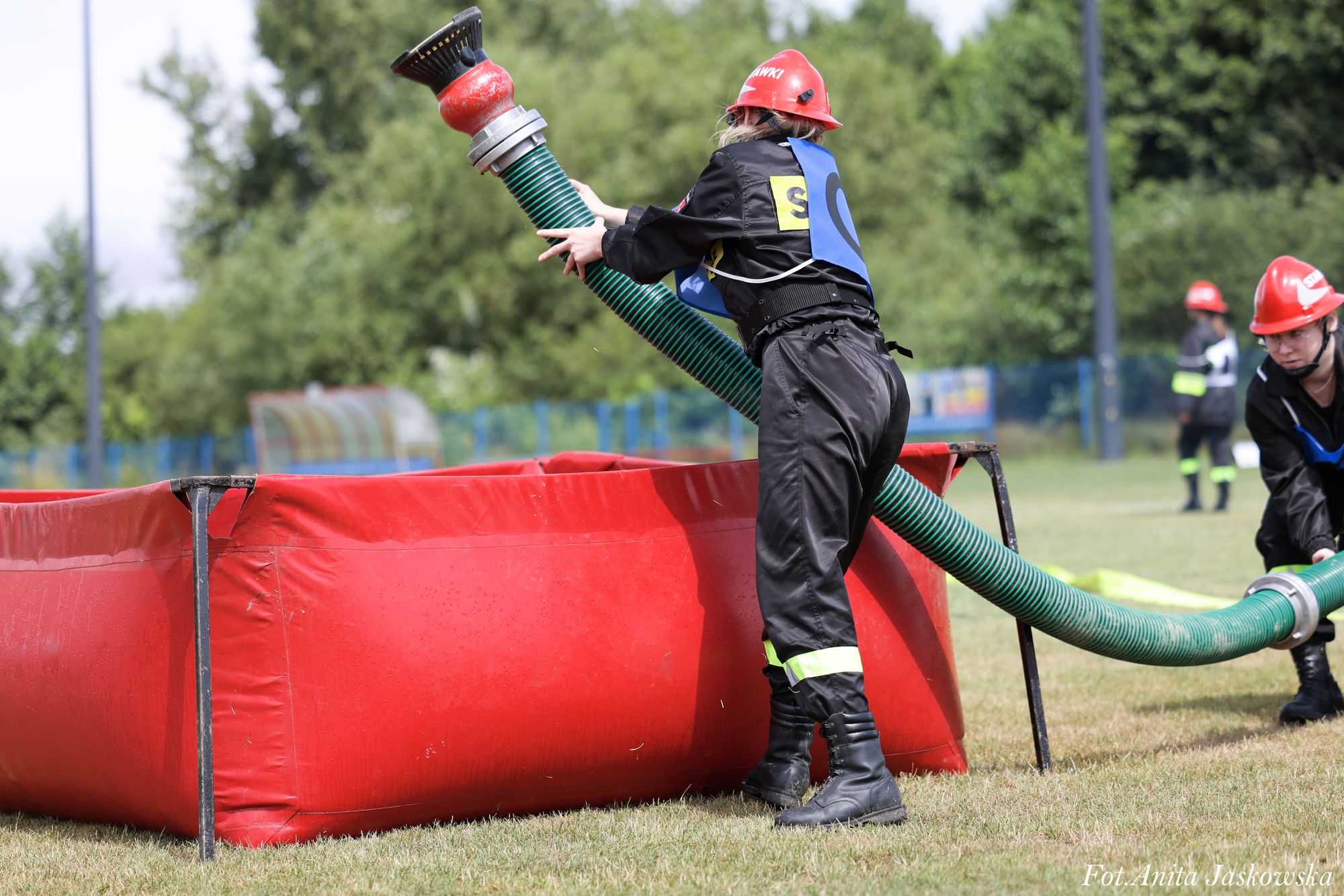 Osoba w czarnym ubraniu ochronnym i czerwonym hełmie trzyma zielony wąż strażacki przy czerwonym zbiorniku na trawie, w tle drzewa i inne osoby w podobnym stroju.
