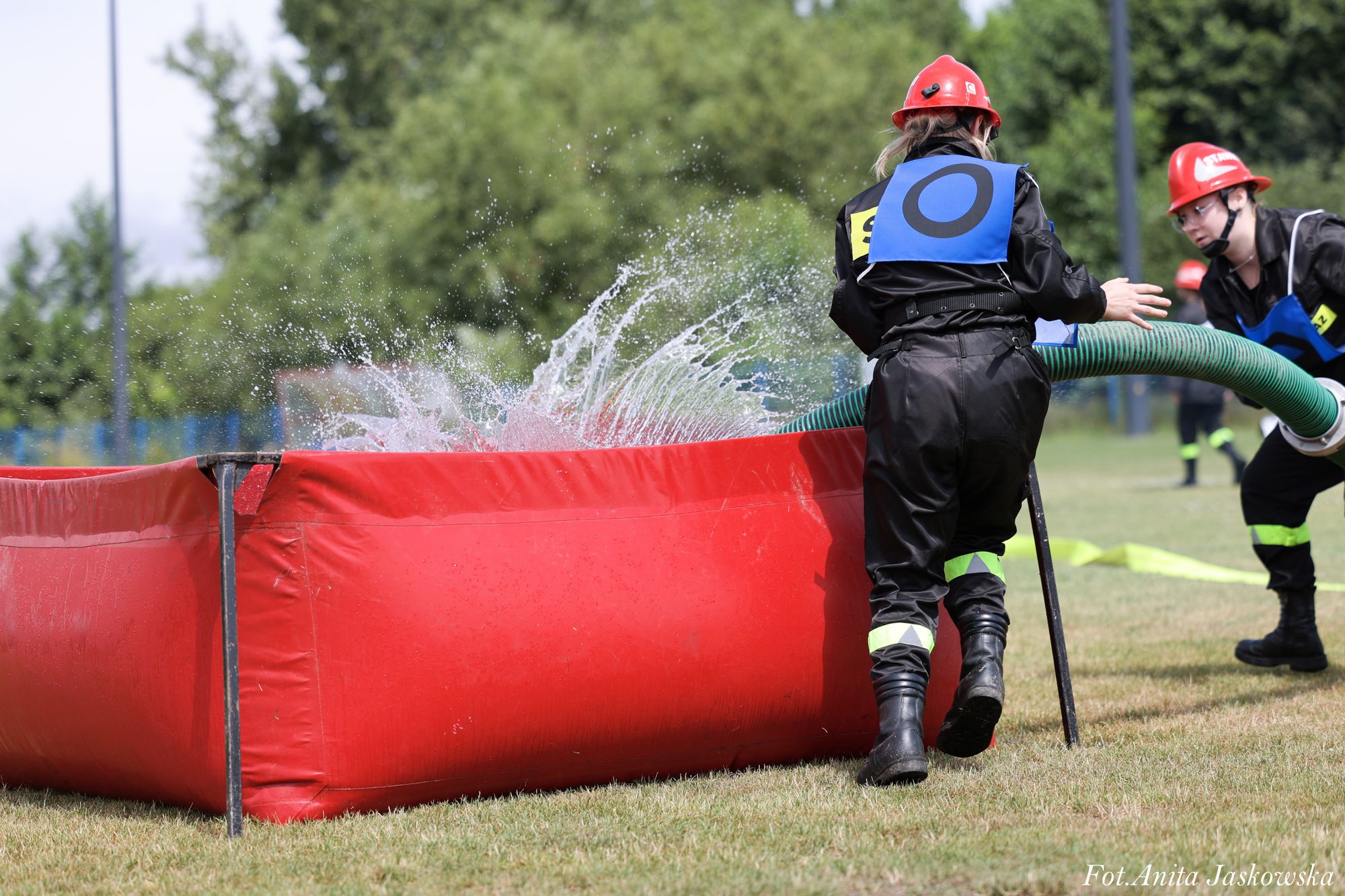 Dwie osoby w czarnych kombinezonach i czerwonych hełmach napełniają zielony wąż wodą czerpaną do dużego czerwonego zbiornika na trawie, w tle zieleń drzew.