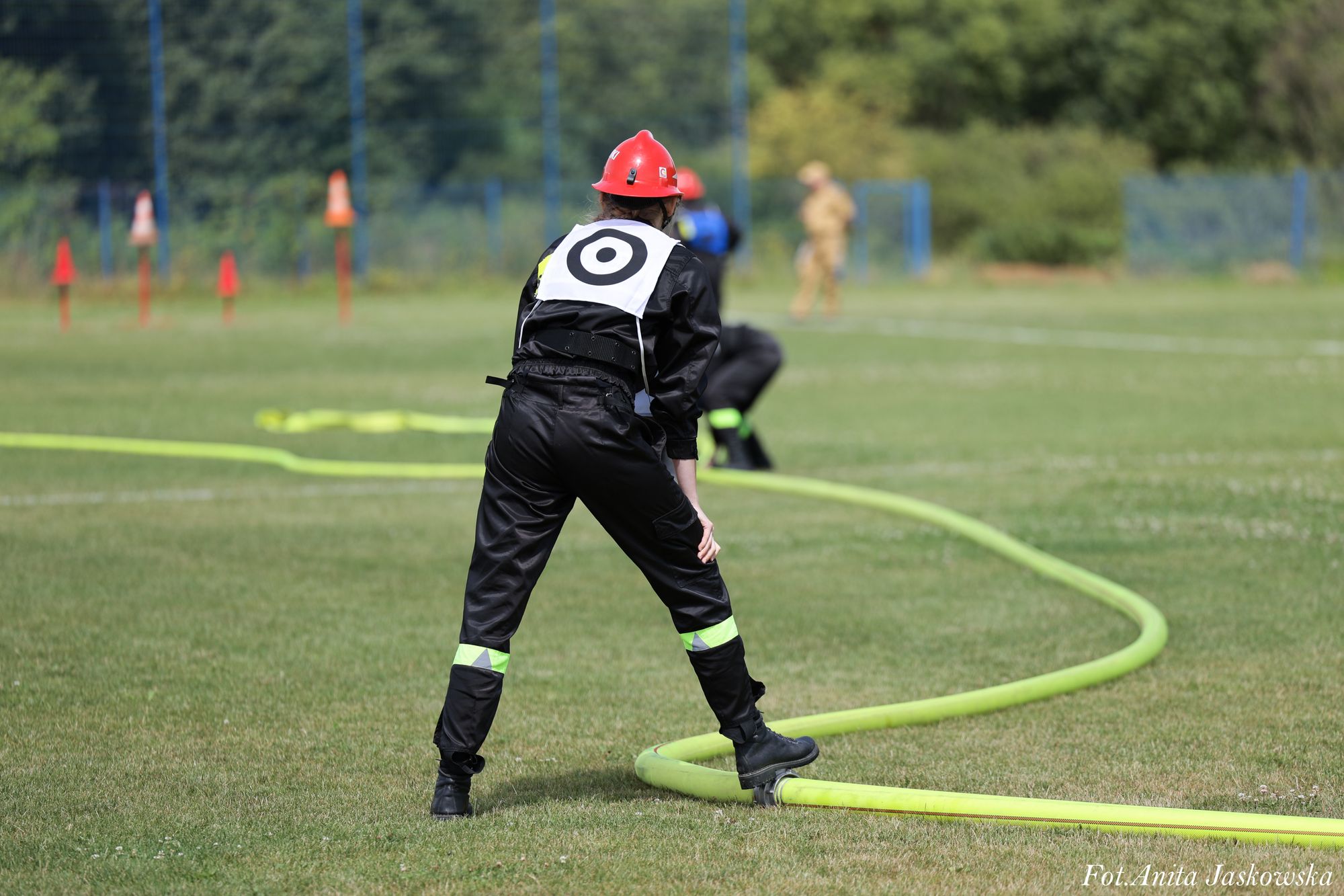 Osoba w czarnym ubraniu z odblaskowymi paskami i czerwonym kaskiem stoi na zielonej trawie, trzymając zielony wąż strażacki, w tle drzewa i niewyraźne sylwetki ludzi.