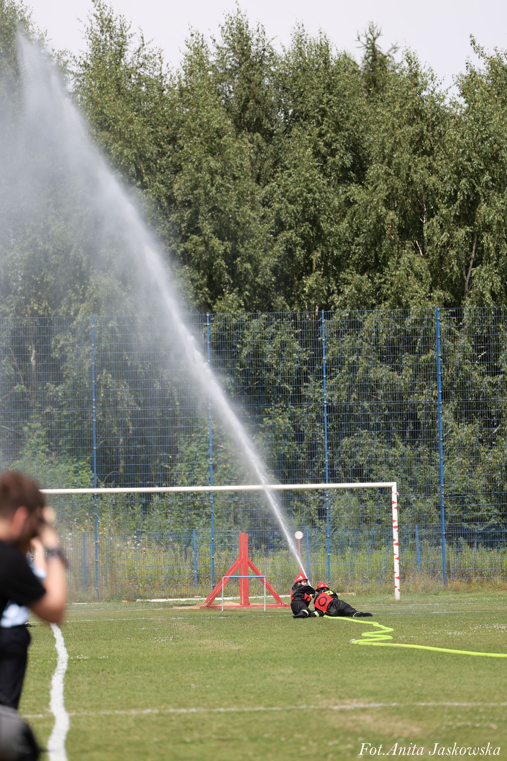 Dwoje dzieci w czarnych strojach i czerwonych kaskach leży na trawie, trzymając strumień wody z węża strażackiego, w tle bramka piłkarska i zielone drzewa.