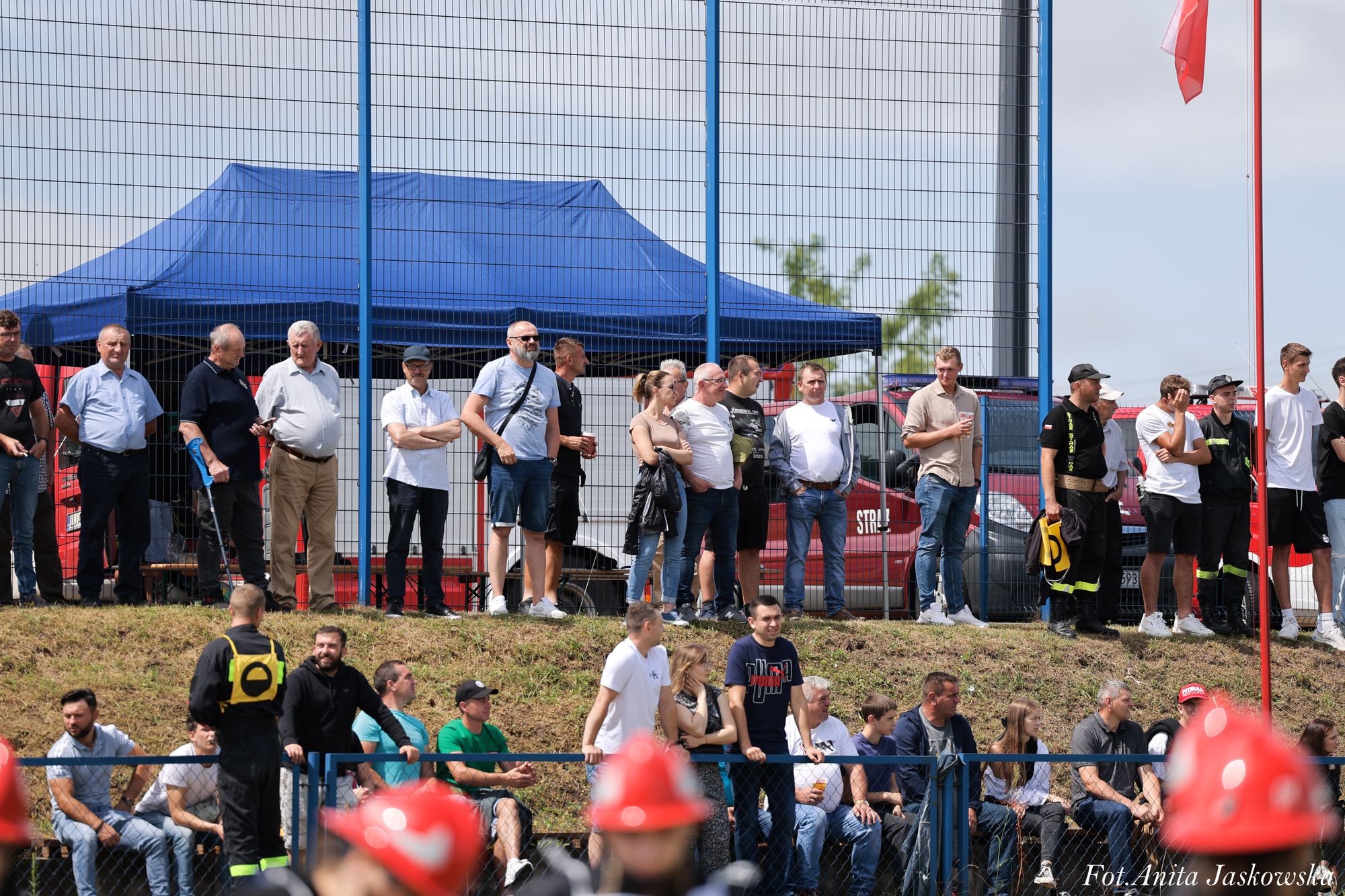 Grupa ludzi stojących i siedzących na skarpie przy ogrodzeniu, w tle niebieski namiot i czerwone pojazdy strażackie, fragmenty czerwonych hełmów strażackich na pierwszym planie.