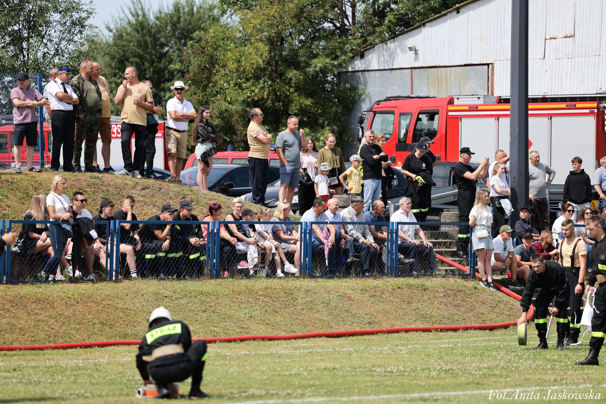 Grupa ludzi stojących i siedzących przy niebieskim ogrodzeniu na tle zielonej trawy, strażacy w czarnych mundurach z żółtymi napisami na polu, czerwony samochód strażacki, drzewa i budynek z blachy falistej.