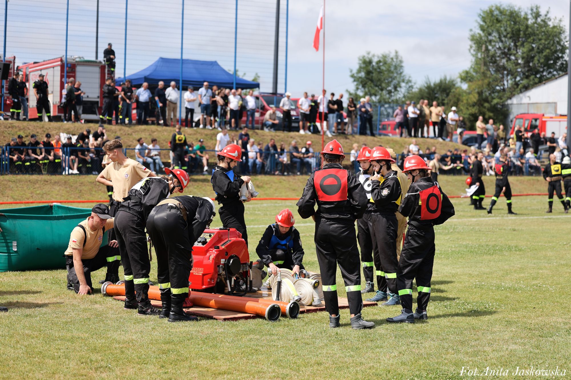 Grupa osób w czarnych mundurach i czerwonych hełmach wokół czerwonego sprzętu strażackiego na trawie, w tle widownia i pojazdy strażackie.