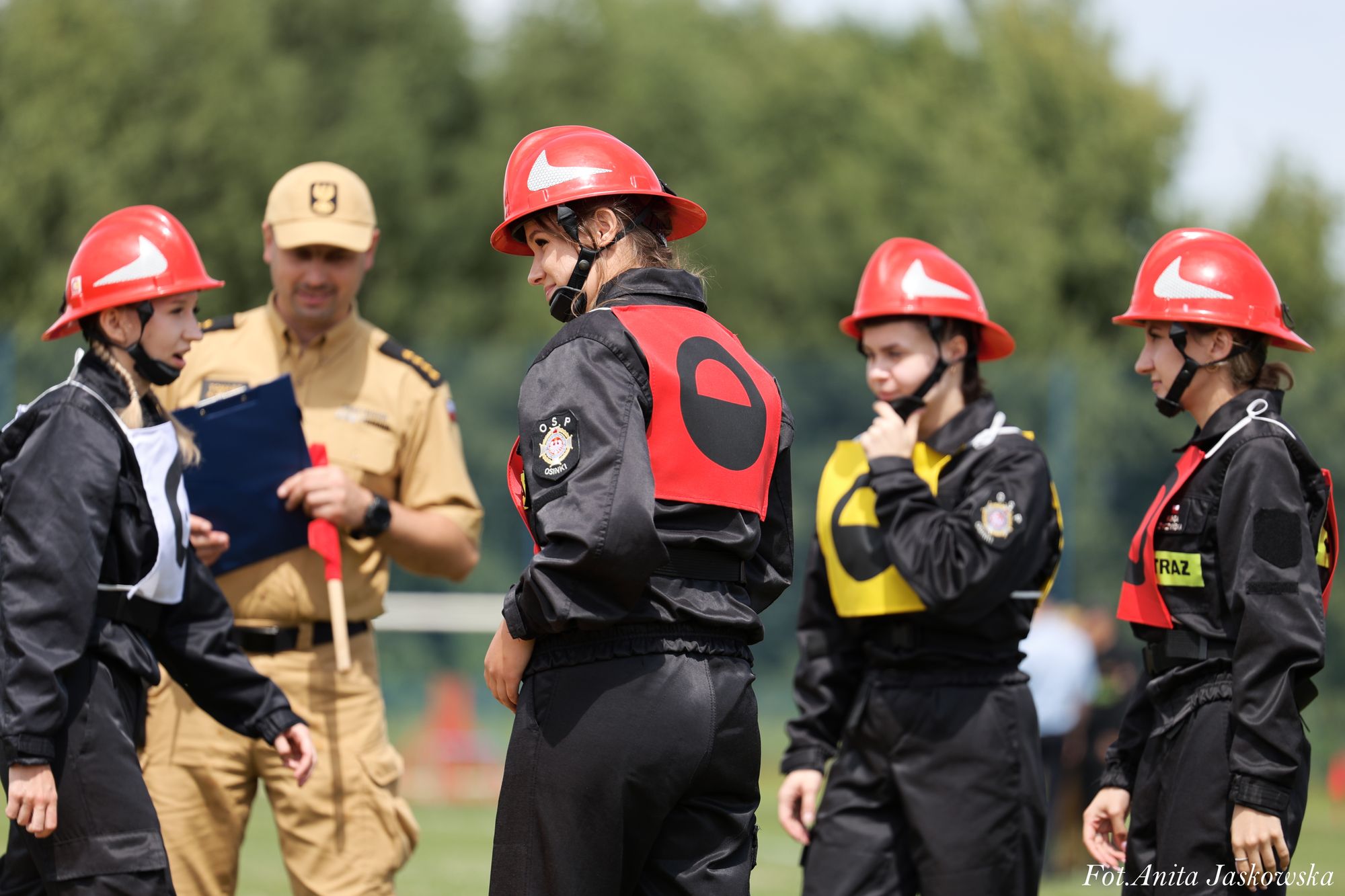 Cztery młode osoby w czarnych uniformach i czerwonych hełmach oraz mężczyzna w beżowym mundurze na tle zieleni. Kolory czerwony, czarny, żółty i beżowy.