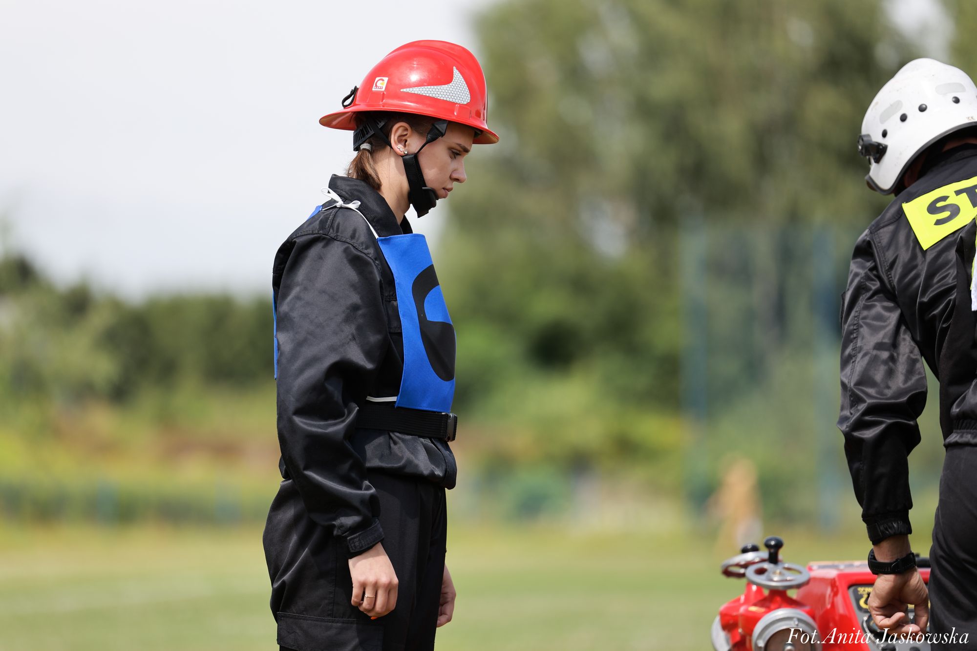 Kobieta w czerwonym kasku i czarnym ubraniu z niebieską kamizelką na tle zieleni, obok mężczyzna w białym kasku i czarnej odzieży, czerwony sprzęt.