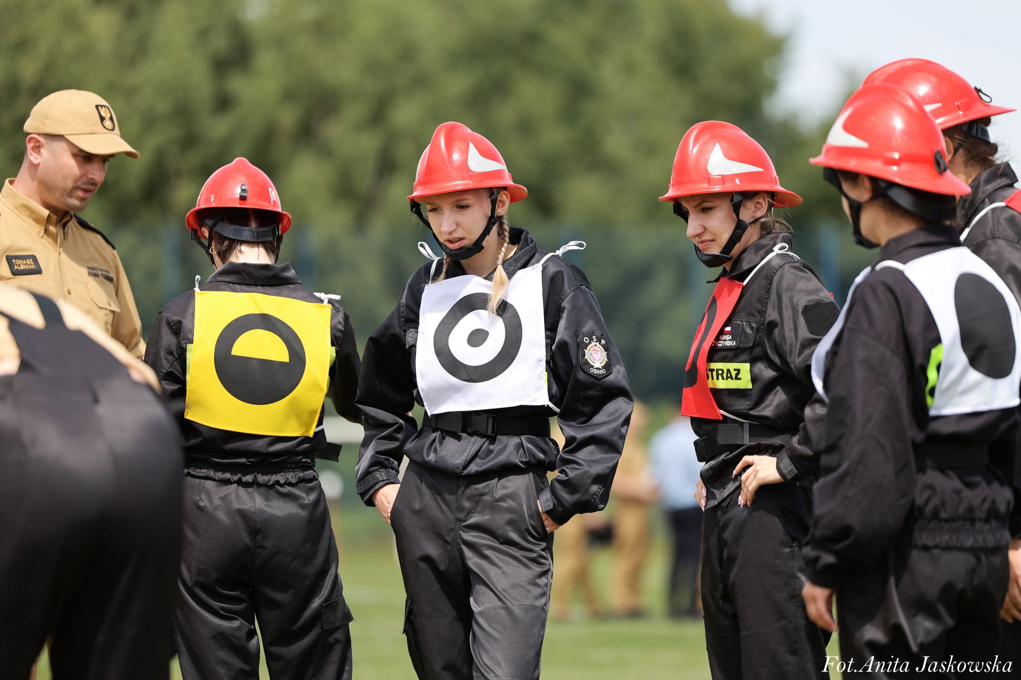 Grupa młodych strażaków w czarnych mundurach i czerwonych hełmach, na koszulach kolorowe oznaczenia, na zewnątrz, zielone tło drzew.