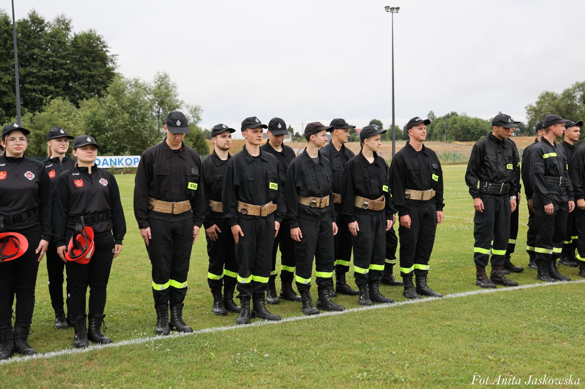 Grupa osób w czarnych mundurach z żółtymi odblaskowymi pasami, stojących na zielonej trawie na tle drzew i nieba, część trzyma czerwone hełmy.