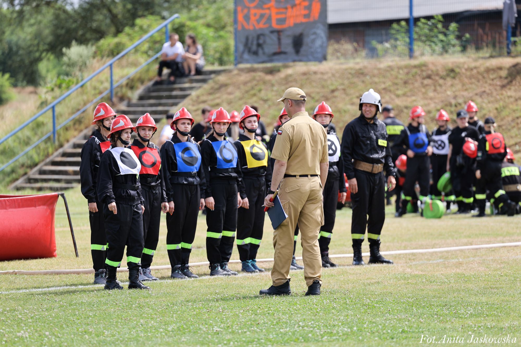 Grupa osób w czarnych mundurach z kolorowymi kamizelkami i czerwonymi hełmami stoi na zielonej trawie, przed nimi stoi mężczyzna w jasnym mundurze z teczką.