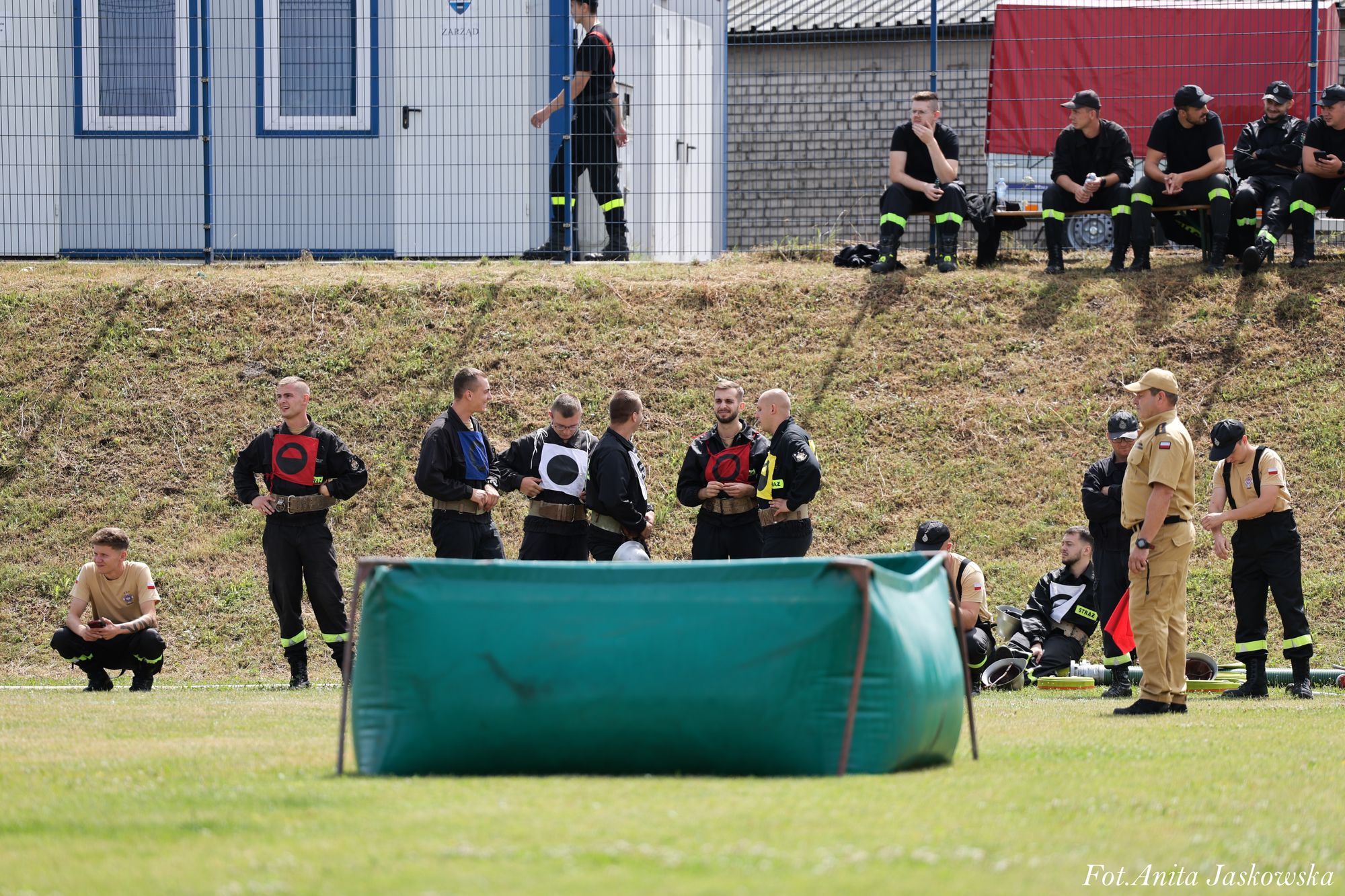 Grupa osób w mundurach strażackich i beżowych ubraniach na trawie, za nimi zielony zbiornik i ogrodzenie z budynkami w tle.