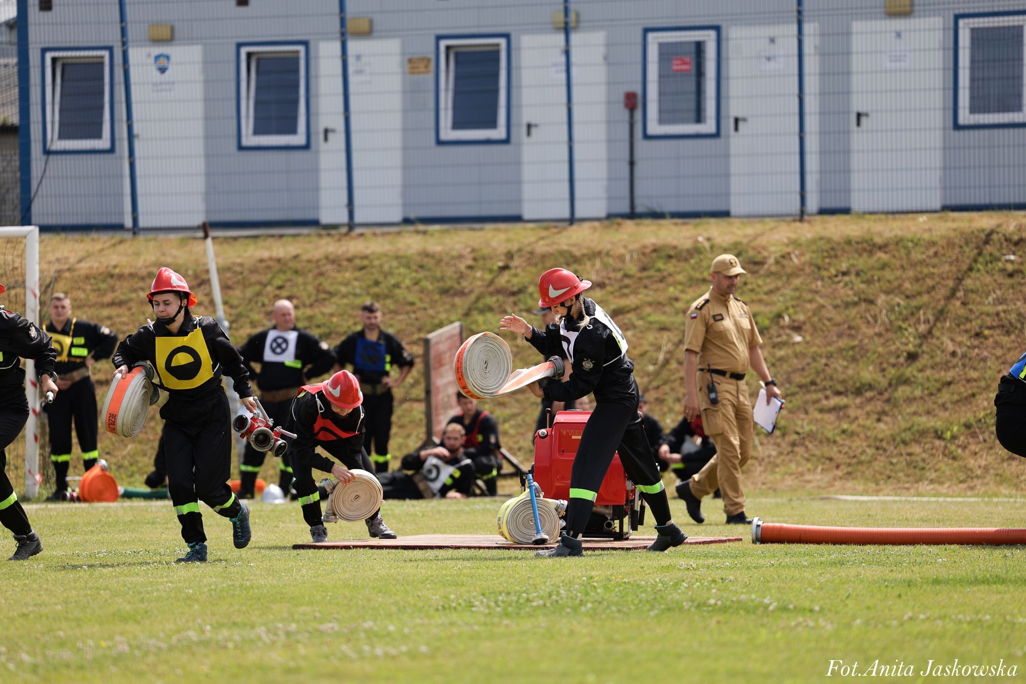 Grupa osób w czarnych strojach z czerwonymi hełmami rozkładających białe i pomarańczowe zwinięte węże pożarnicze na zielonym boisku, w tle budynki i obserwujący mężczyzna w beżowym mundurze.