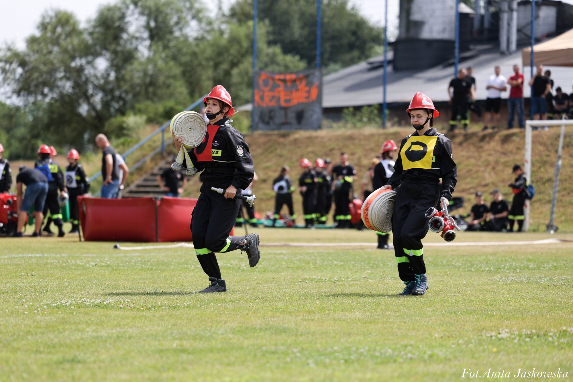 Dwóch strażaków w czarnych mundurach i czerwonych hełmach niesie zwinięte białe węże strażackie na zielonym boisku, w tle ludzie i napis OSP Krzemień.