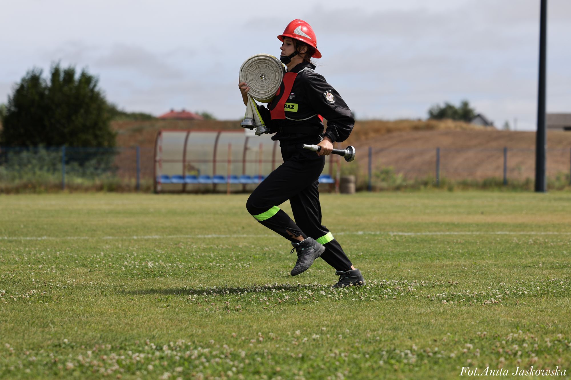 Strażak w czarnym ubraniu z odblaskowymi paskami i czerwonym hełmem biegnie po trawie, niosąc zwinięty wąż strażacki i metalową końcówkę.