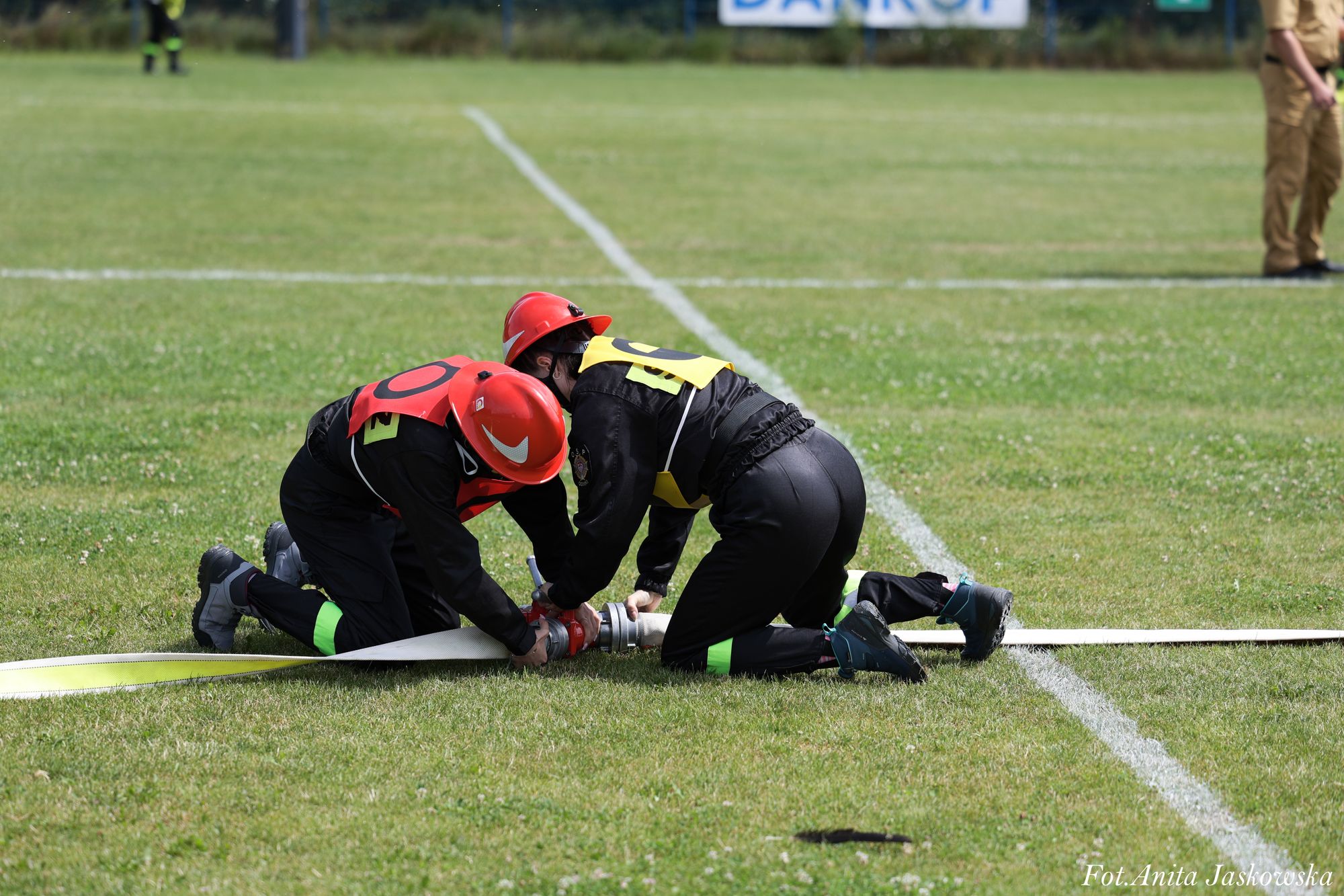 Dwóch strażaków w czerwonych hełmach i ubraniach ochronnych klęczy na zielonej trawie, łącząc metalowe końcówki białego węża strażackiego.
