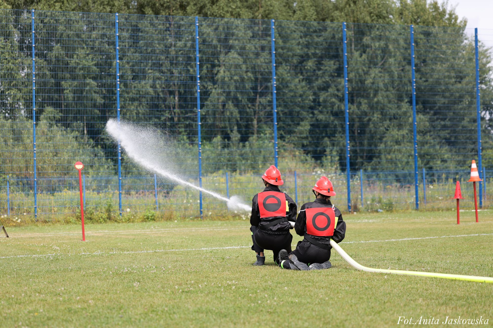 Dwie osoby w czarnych uniformach i czerwonych kaskach klęczą na trawie, jedna trzyma wąż strażacki, z którego leci strumień wody, w tle niebieski płot i drzewa.
