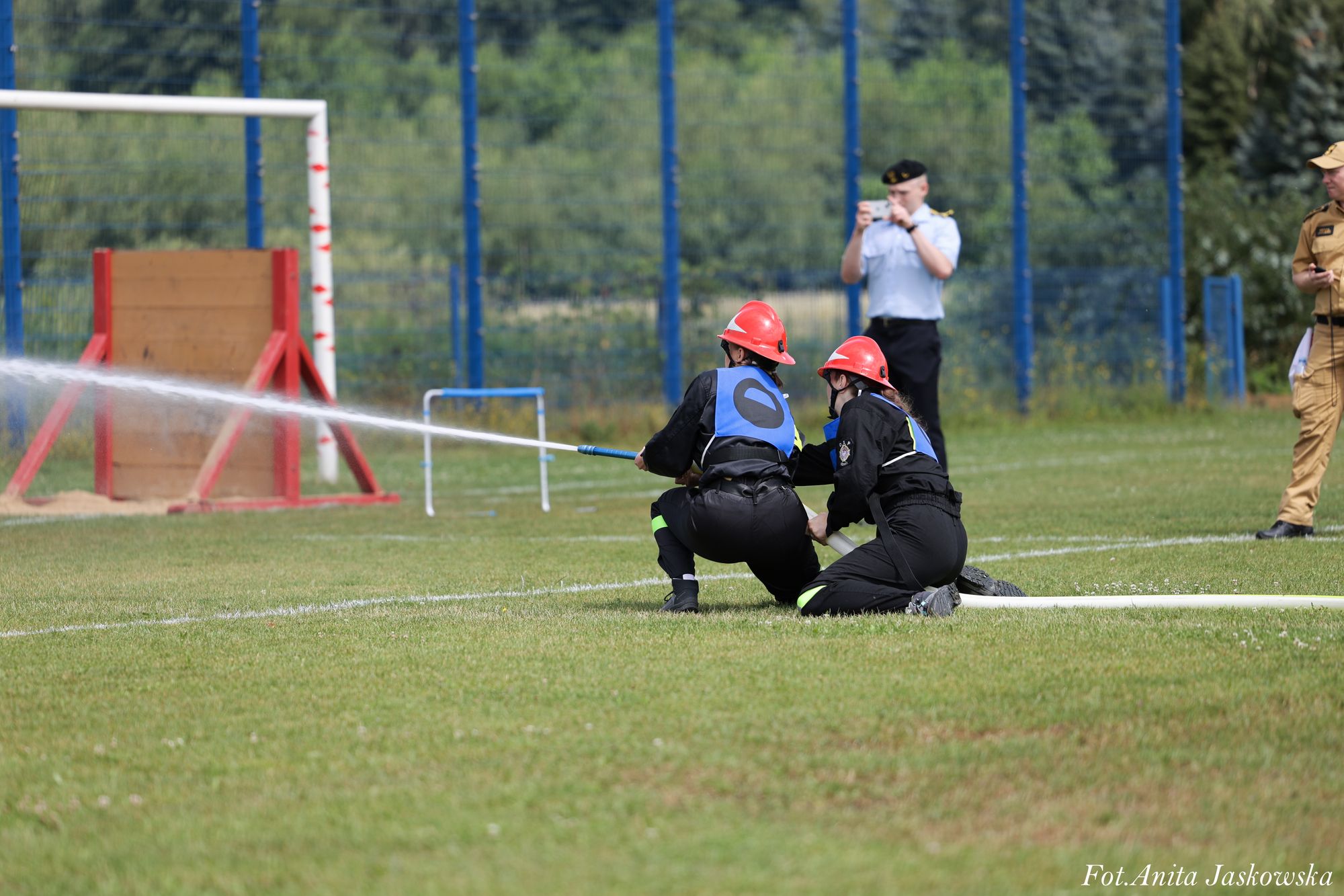 Dwóch strażaków w czarnych mundurach i czerwonych hełmach obsługuje prądownicę wodną na zielonym boisku, w tle mężczyzna robiący zdjęcie i inny w mundurze beżowym.