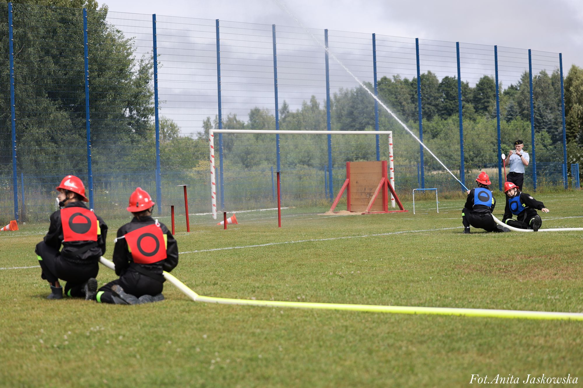 Cztery osoby w czarnych strojach i czerwonych hełmach, dwie z czerwonymi i dwie z niebieskimi kamizelkami, trzymają węże strażackie na zielonym boisku, w tle niebieski ogrodzenie i drzewa.