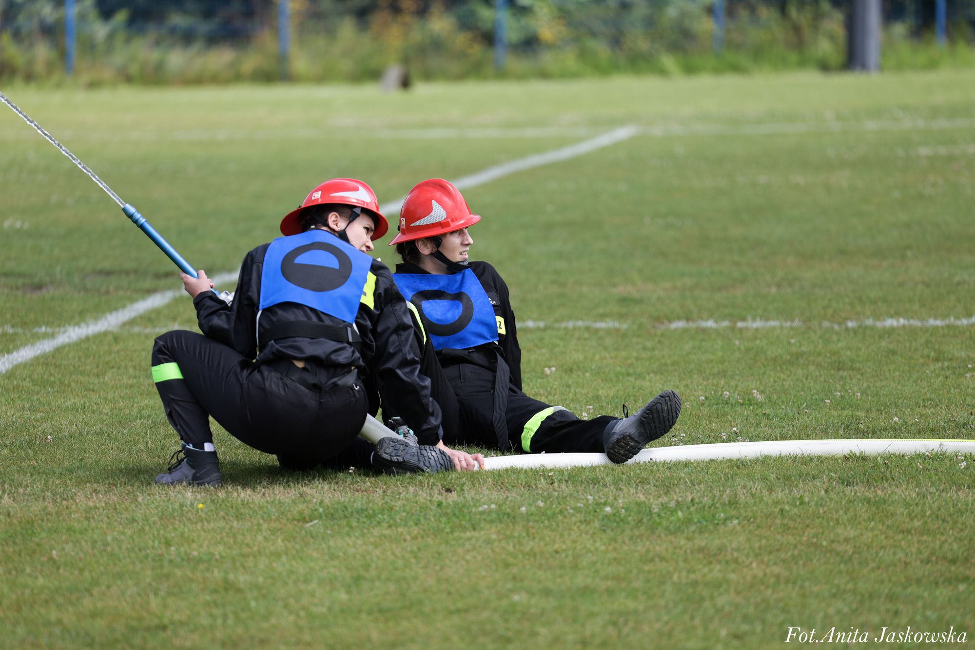 Dwie osoby w czarnych ubraniach i czerwonych hełmach siedzą na zielonej trawie, trzymając biały wąż strażacki z niebieską końcówką, z której leci strumień wody.