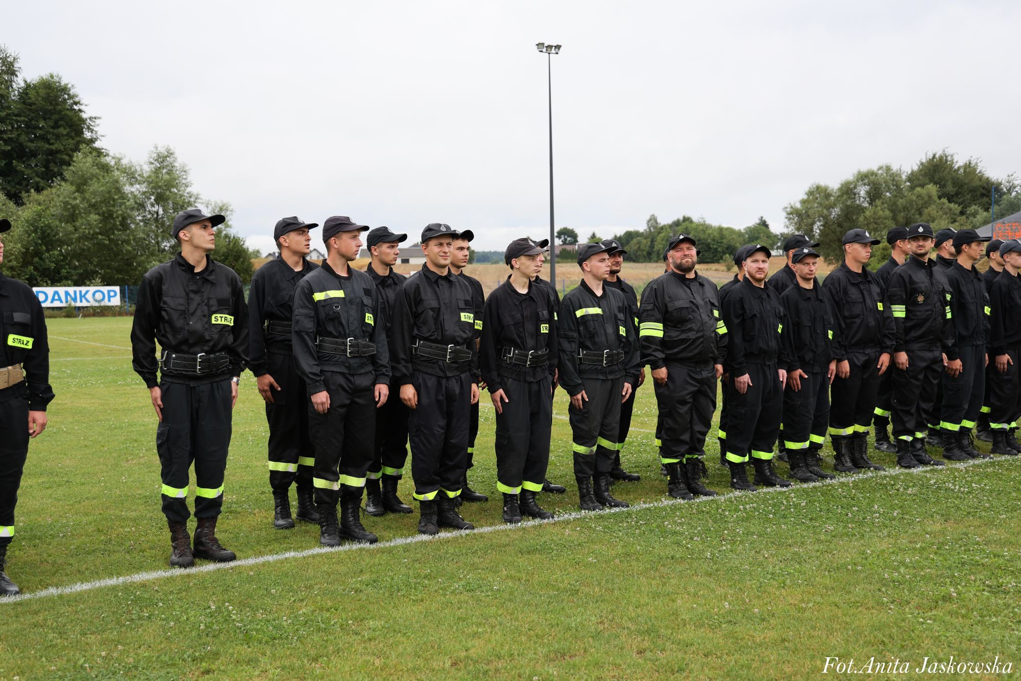 Grupa mężczyzn w czarnych mundurach z żółtymi odblaskowymi naszywkami i paskami, stojących na zielonej trawie na tle drzew i nieba.