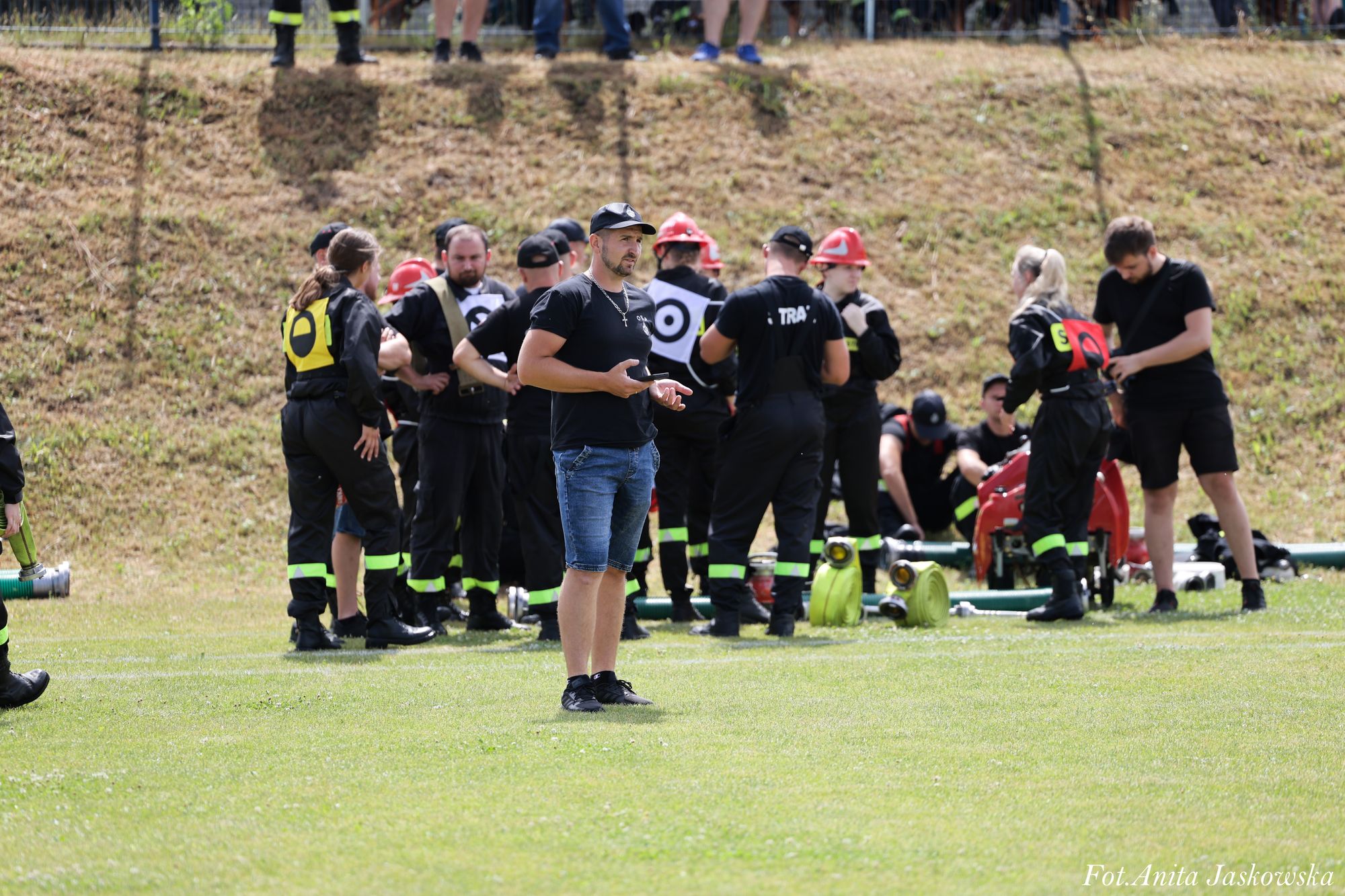 Grupa osób w czarnych ubraniach z neonowymi odblaskami i czerwonymi hełmami na trawie, mężczyzna w czarnej koszulce i niebieskich spodenkach na pierwszym planie.