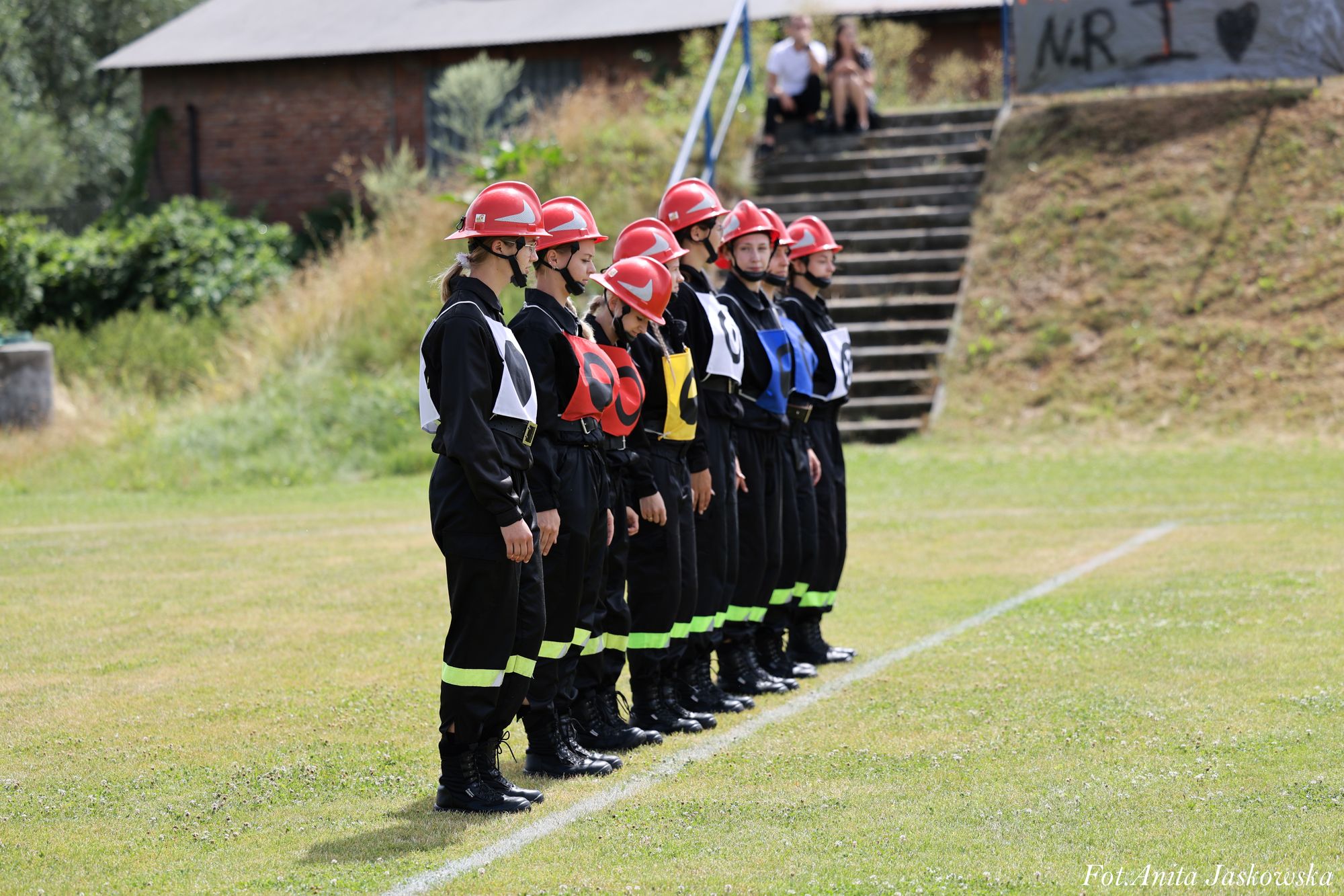 Siedem osób w czarnych mundurach z odblaskowymi paskami, czerwonych hełmach i kolorowych numerach na piersiach stoi na zielonym boisku obok białej linii.