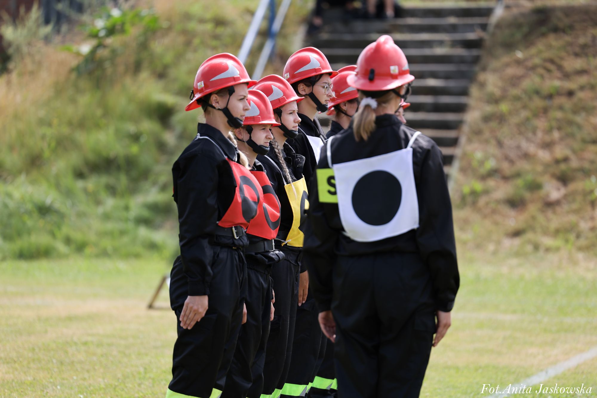 Grupa osób w czarnych mundurach i czerwonych hełmach stoi w szeregu na trawie, każda z kolorową tabliczką na piersi, w tle schody i zarośla.