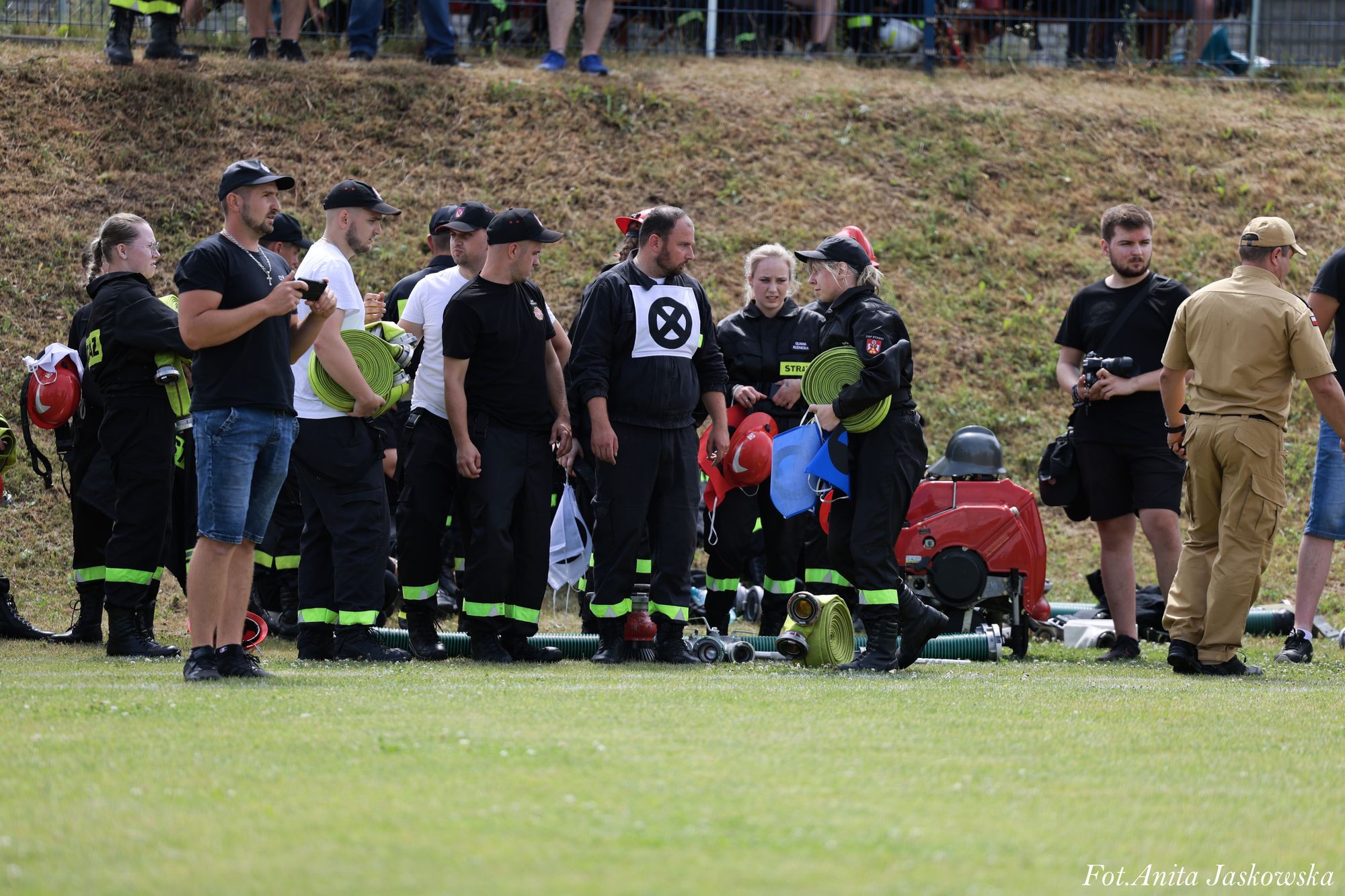 Grupa ludzi w mundurach strażackich i cywilnych stoi na trawie, niektórzy trzymają zwinięte zielone węże strażackie i czerwone hełmy, w tle teren lekko pagórkowaty.