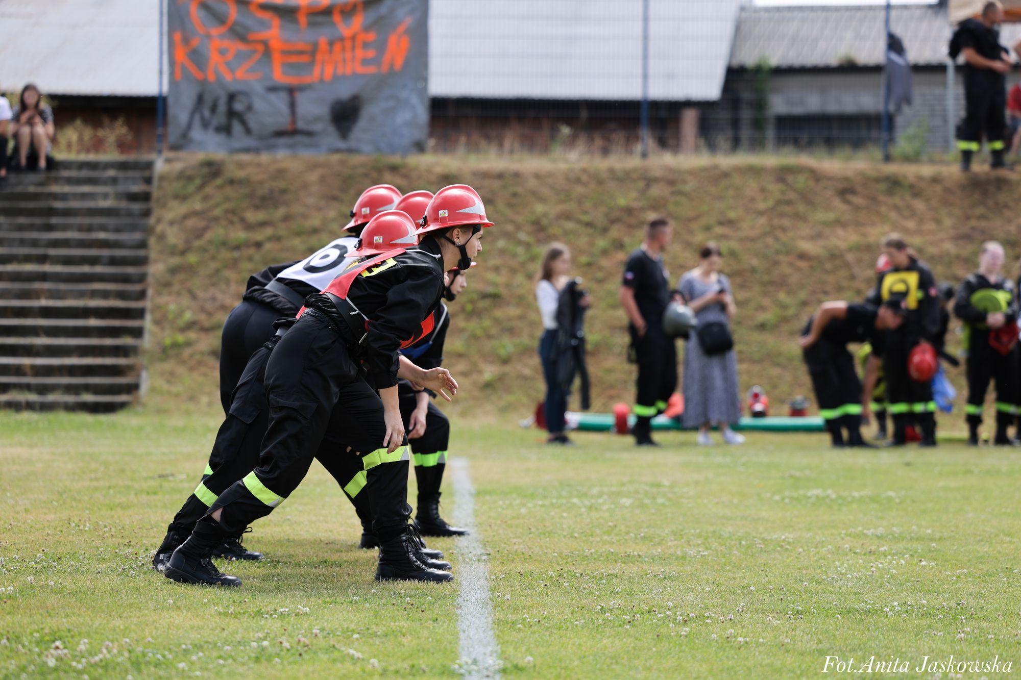 Cztery osoby w czarnych ubraniach z żółtymi odblaskami i czerwonych hełmach pochylone na linii startu na zielonej trawie, w tle grupa ludzi i transparent z napisem.
