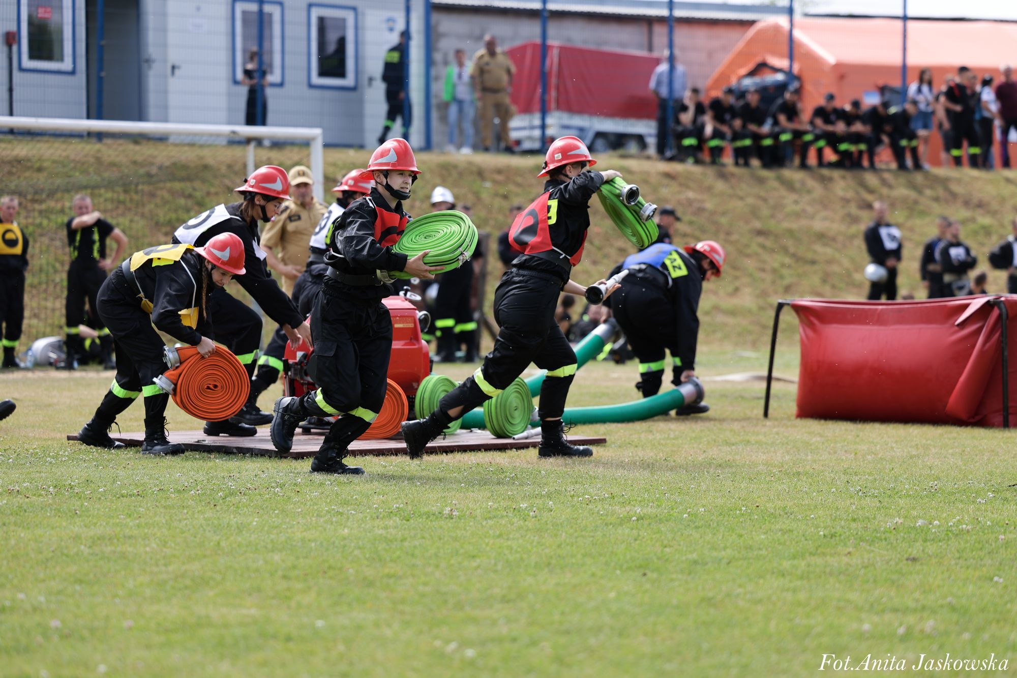 Grupa osób w czarnych kombinezonach i czerwonych hełmach trzyma zielone i pomarańczowe zwinięte węże strażackie na trawiastym boisku, w tle widoczni ludzie i budynki.