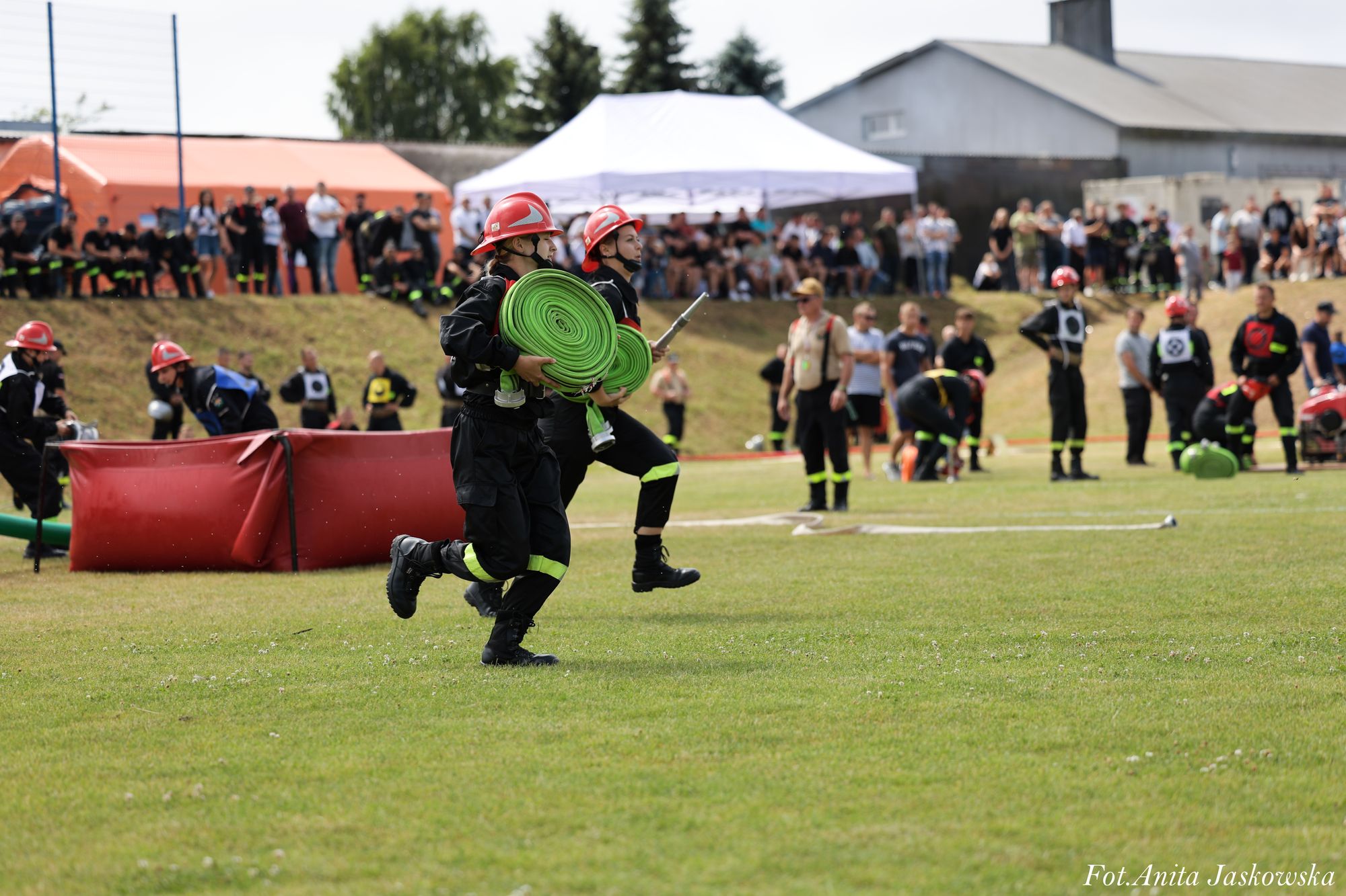 Dwóch strażaków w czerwonych hełmach i czarnych mundurach biegnie trzymając zielone zwinięte węże strażackie na tle tłumu ludzi i namiotu.
