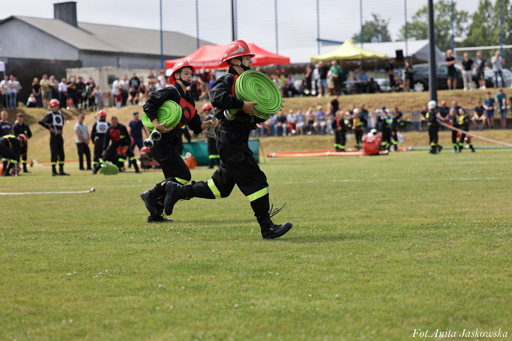 Dwóch strażaków w czarnych mundurach i czerwonych hełmach biegnie trzymając zielone zwinięte węże strażackie, na tle trawiastego boiska i widowni.