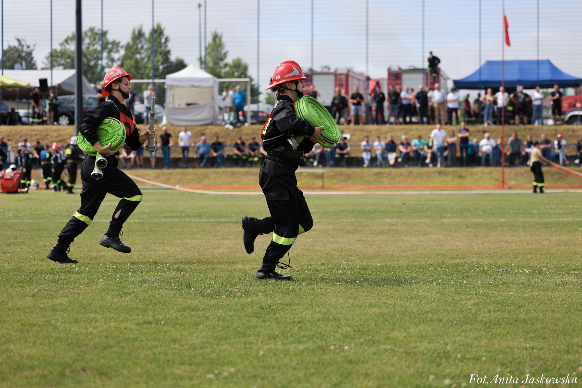 Dwóch strażaków w czarnych mundurach z żółtymi odblaskami i czerwonych hełmach biegnie po zielonym boisku, trzymając zwinięte zielone węże strażackie, w tle widoczna publiczność.