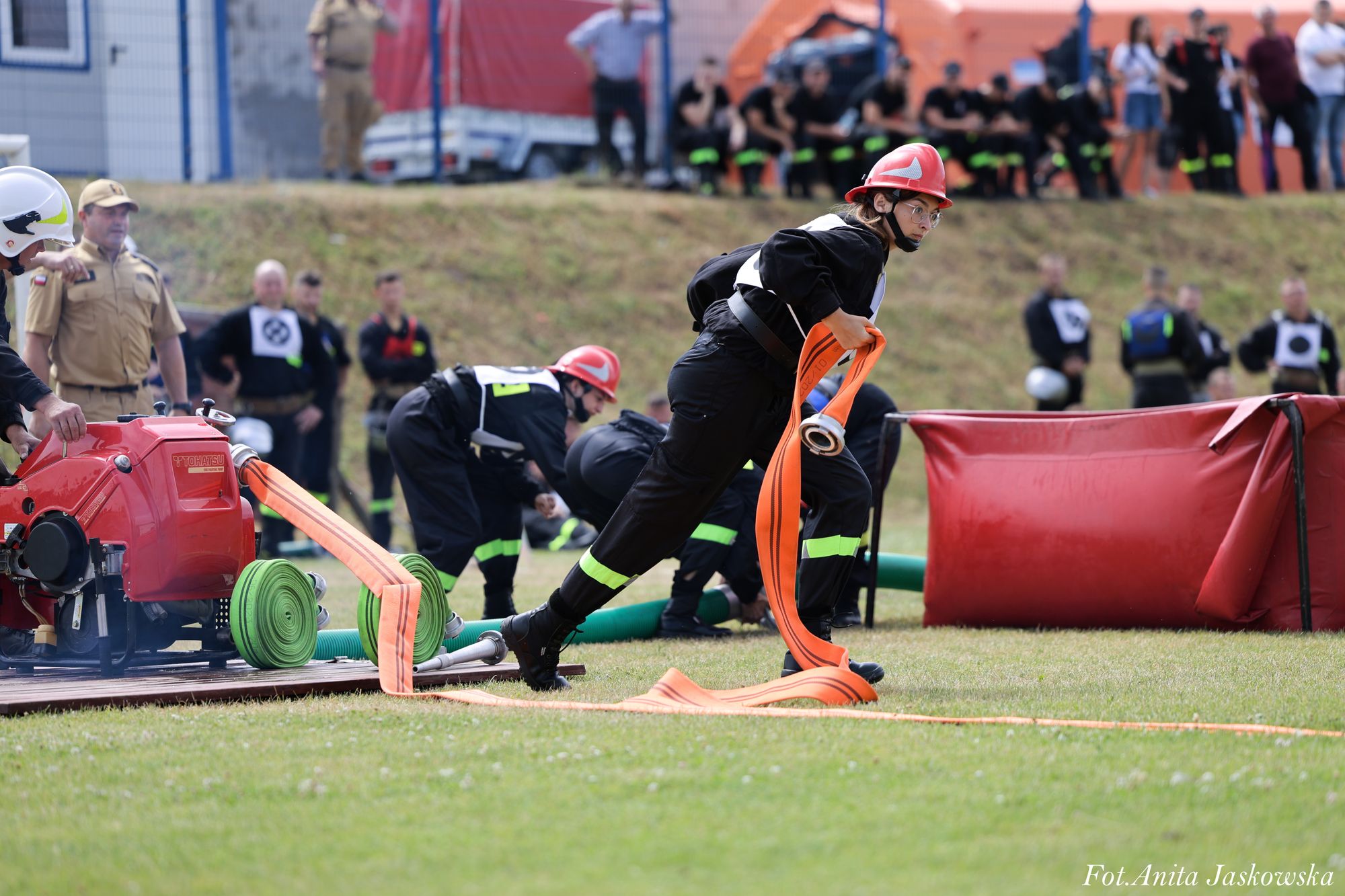 Strażak w czarnym ubraniu ochronnym z czerwonym hełmem rozwija pomarańczowy wąż obok czerwonej pompy na trawie, w tle osoby i sprzęt strażacki.