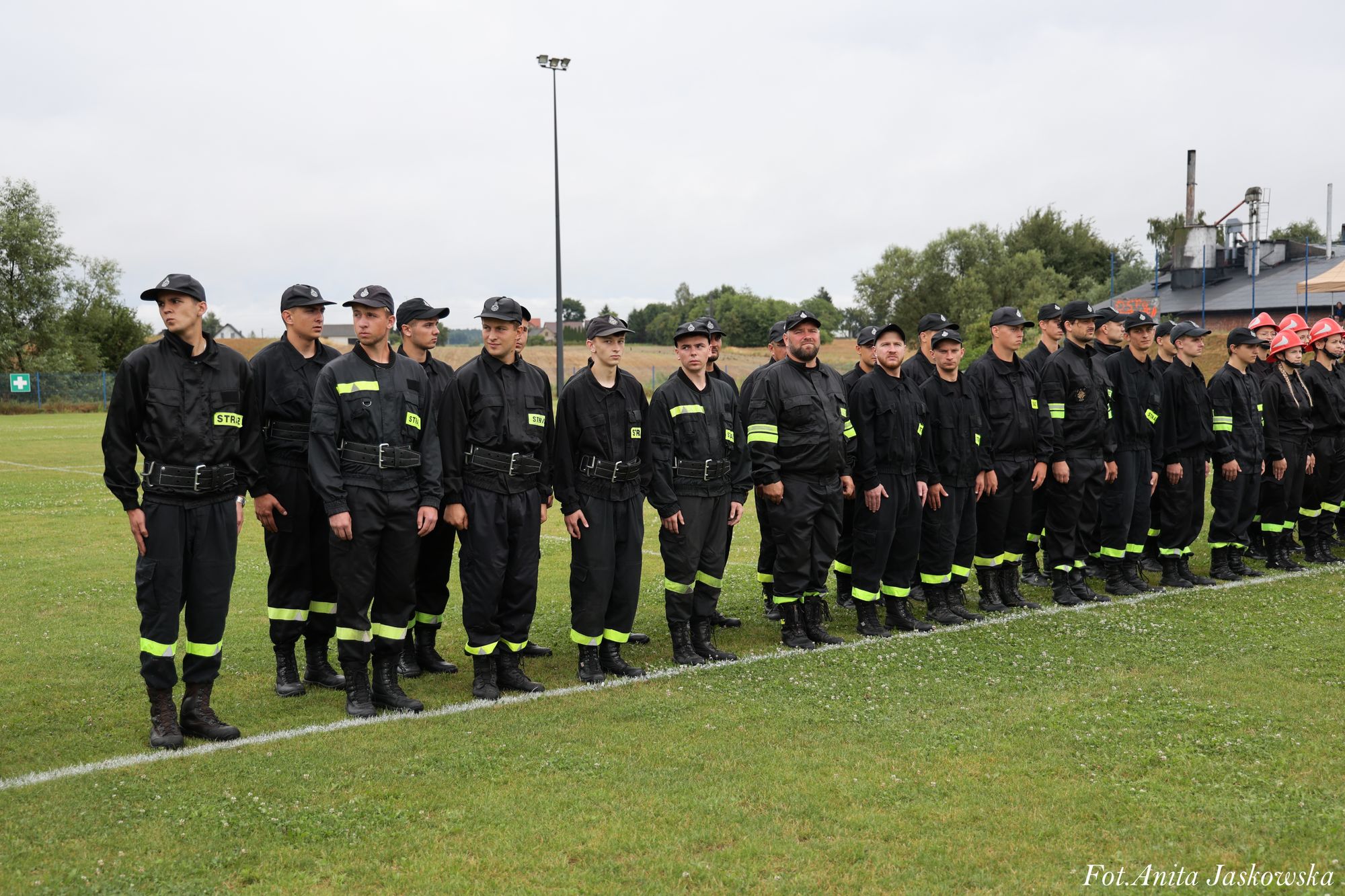 Grupa osób w czarnych mundurach z odblaskowymi żółtymi elementami stoi w szeregu na zielonym boisku, w tle drzewa i budynki.