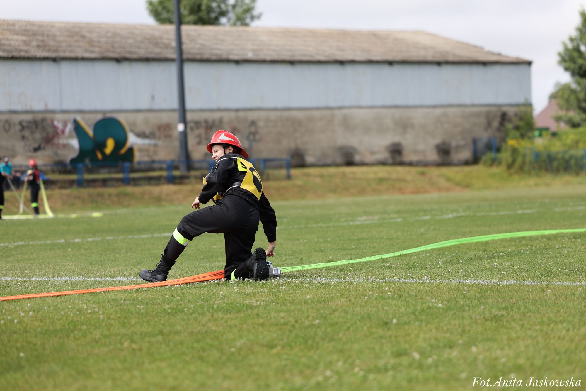 Młoda osoba w czarnym ubraniu i czerwonym kasku na zielonym boisku, trzymająca kolorowe węże strażackie, w tle budynek z graffiti i inni uczestnicy.