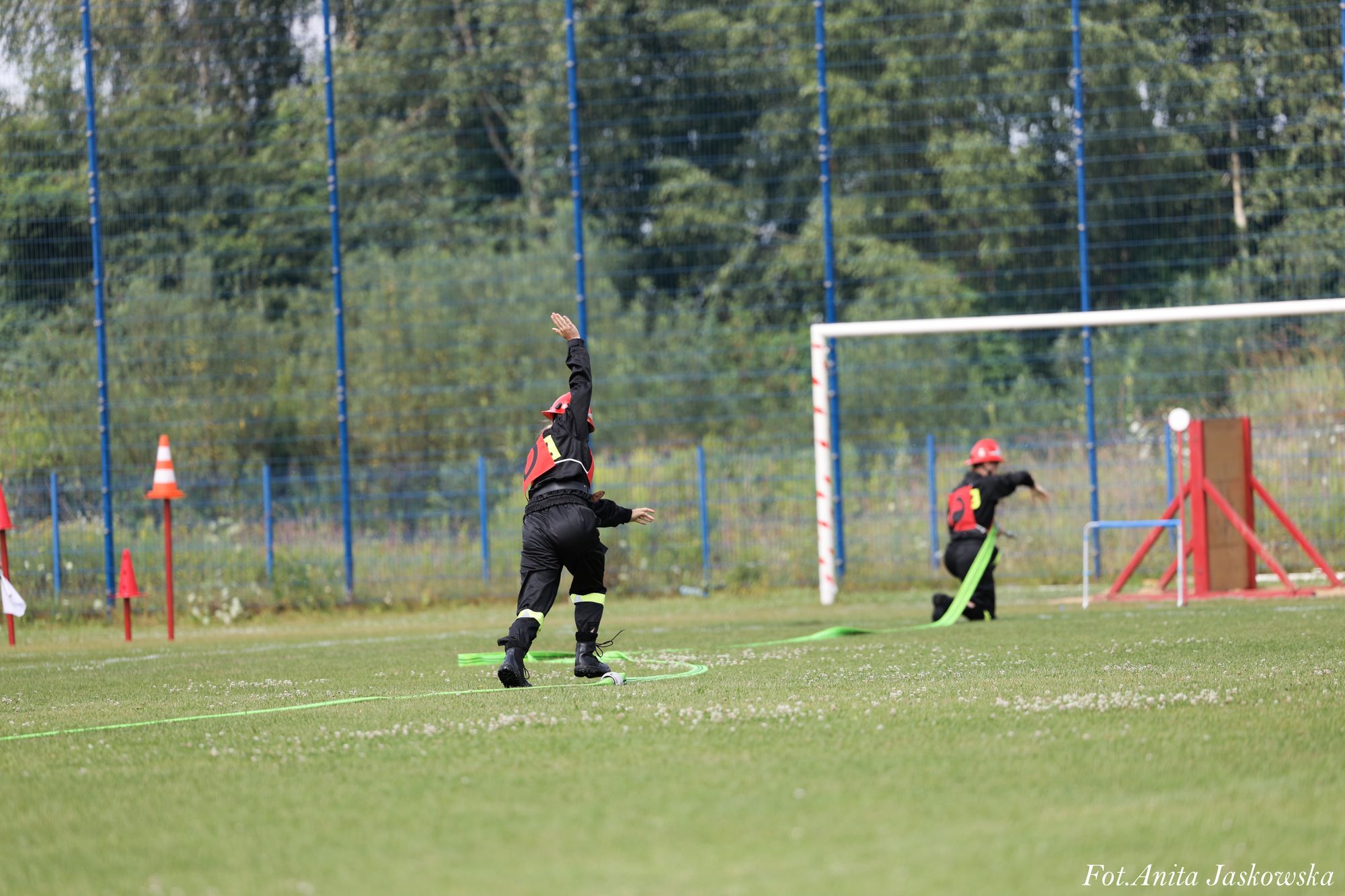 Dwie osoby w czarnych uniformach i czerwonych kaskach na zielonym boisku, jedna stoi z podniesioną ręką, druga klęczy z rozwiniętym zielonym wężem strażackim.