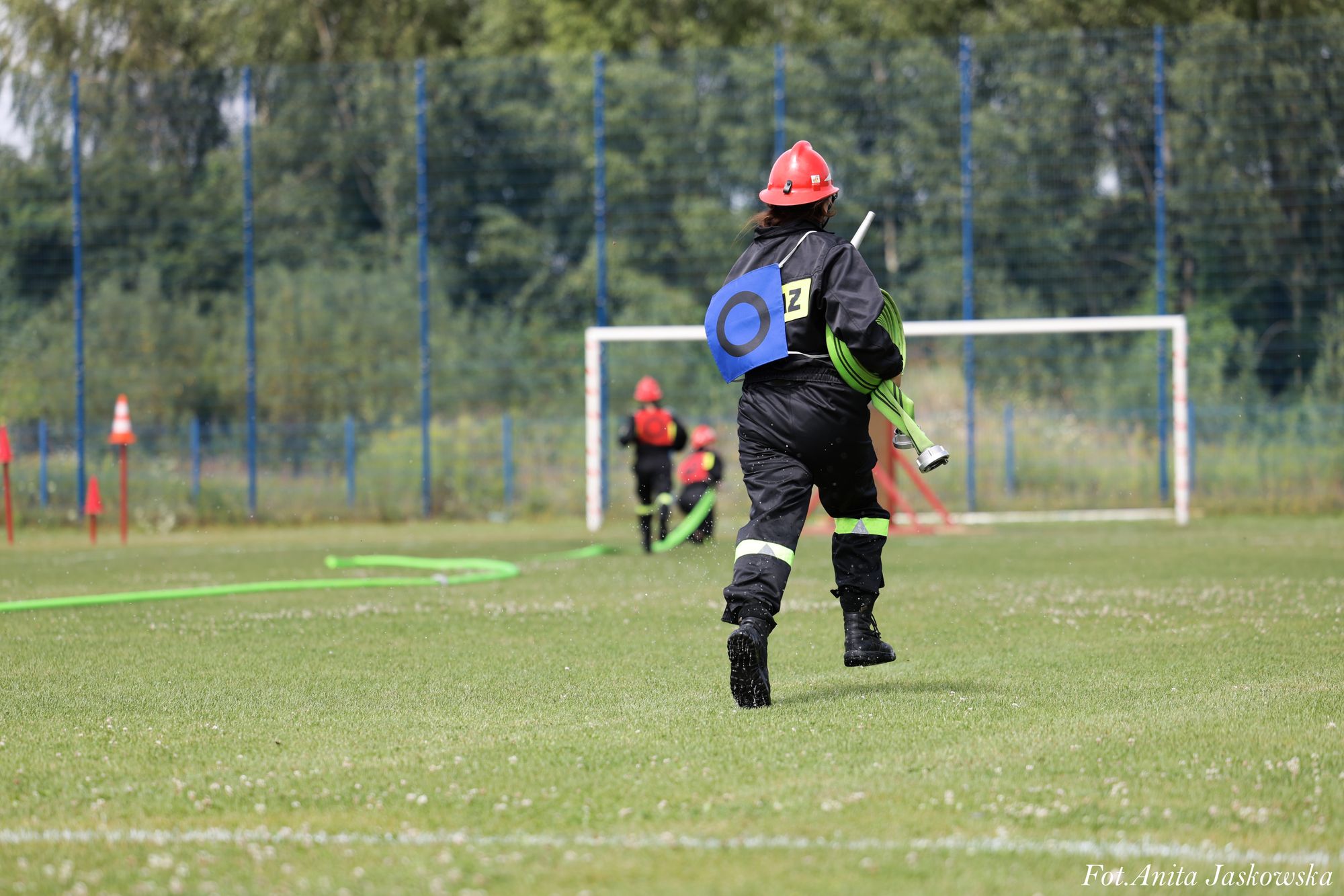 Strażak w czerwonym hełmie i czarnym ubraniu z odblaskowymi pasami, niosący zielony zwinięty wąż strażacki na zielonym boisku sportowym.