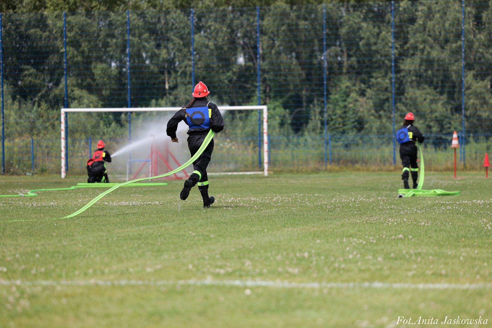 Cztery osoby w czarnych uniformach i czerwonych hełmach trzymają zielone węże strażackie na zielonym boisku sportowym, w tle bramka i siatka.
