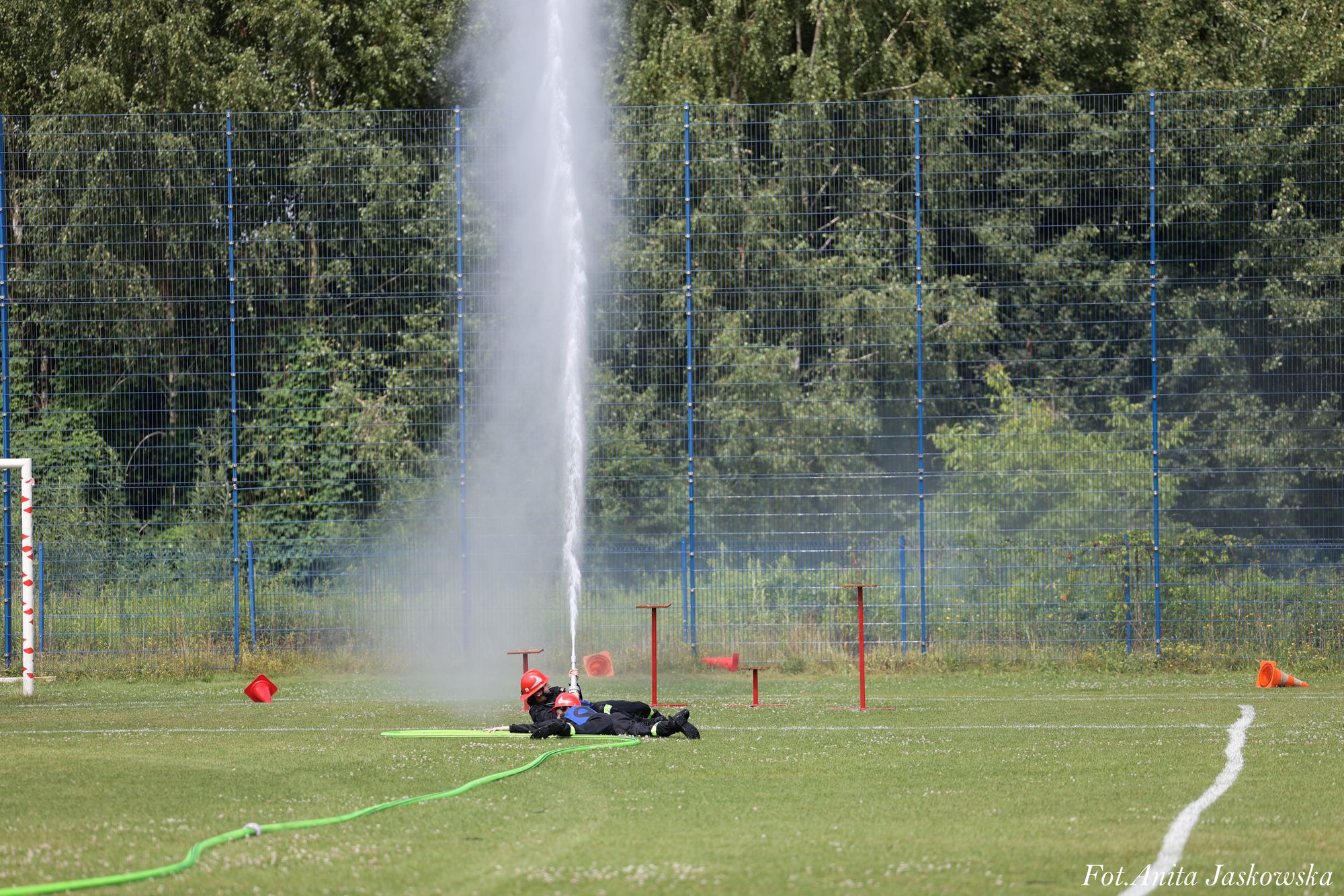 Dwóch strażaków w czarnych mundurach i czerwonych hełmach leży na zielonej trawie, trzymając strumień wody z węża, z tyłu niebieskie ogrodzenie i drzewa.