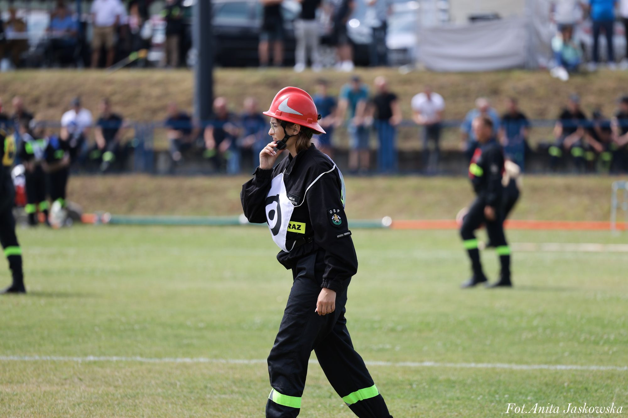Osoba w czarnym ubraniu ochronnym z żółtymi odblaskowymi paskami i czerwonym hełmem na zielonym trawiastym boisku, w tle rozmazani ludzie.