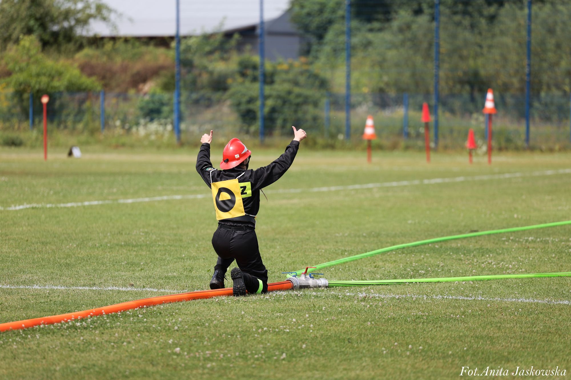 Osoba w czarnym ubraniu i czerwonym hełmie klęczy na trawie obok pomarańczowego i zielonych węży strażackich na boisku sportowym z oznaczeniami.