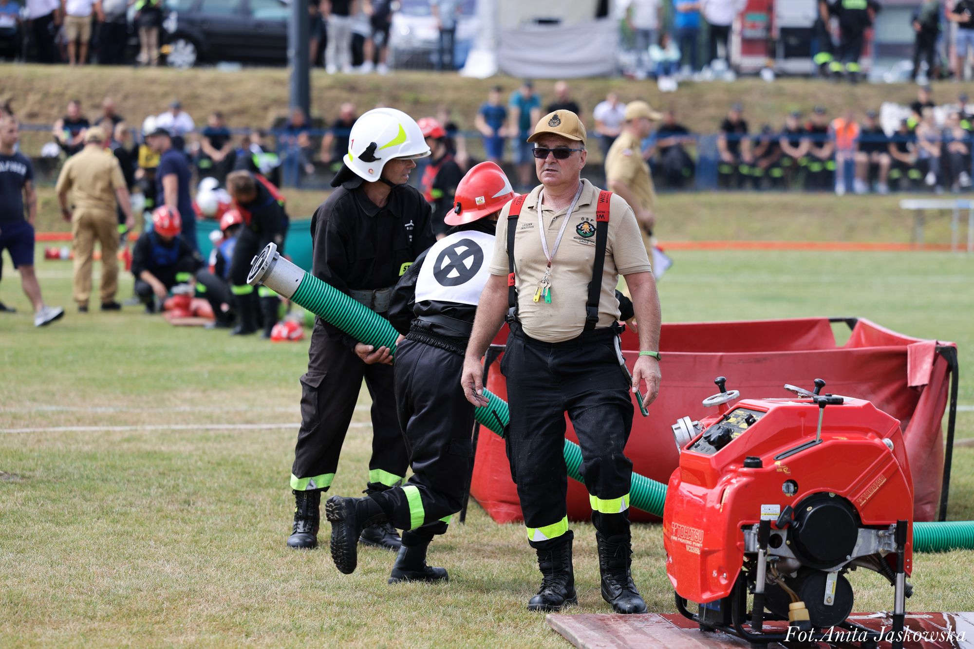 Mężczyzna w beżowej koszulce, czarnych spodniach z żółtymi odblaskami i czapce na tle czerwonej pompy wodnej, w tle inni strażacy w kaskach i zielona trawa.