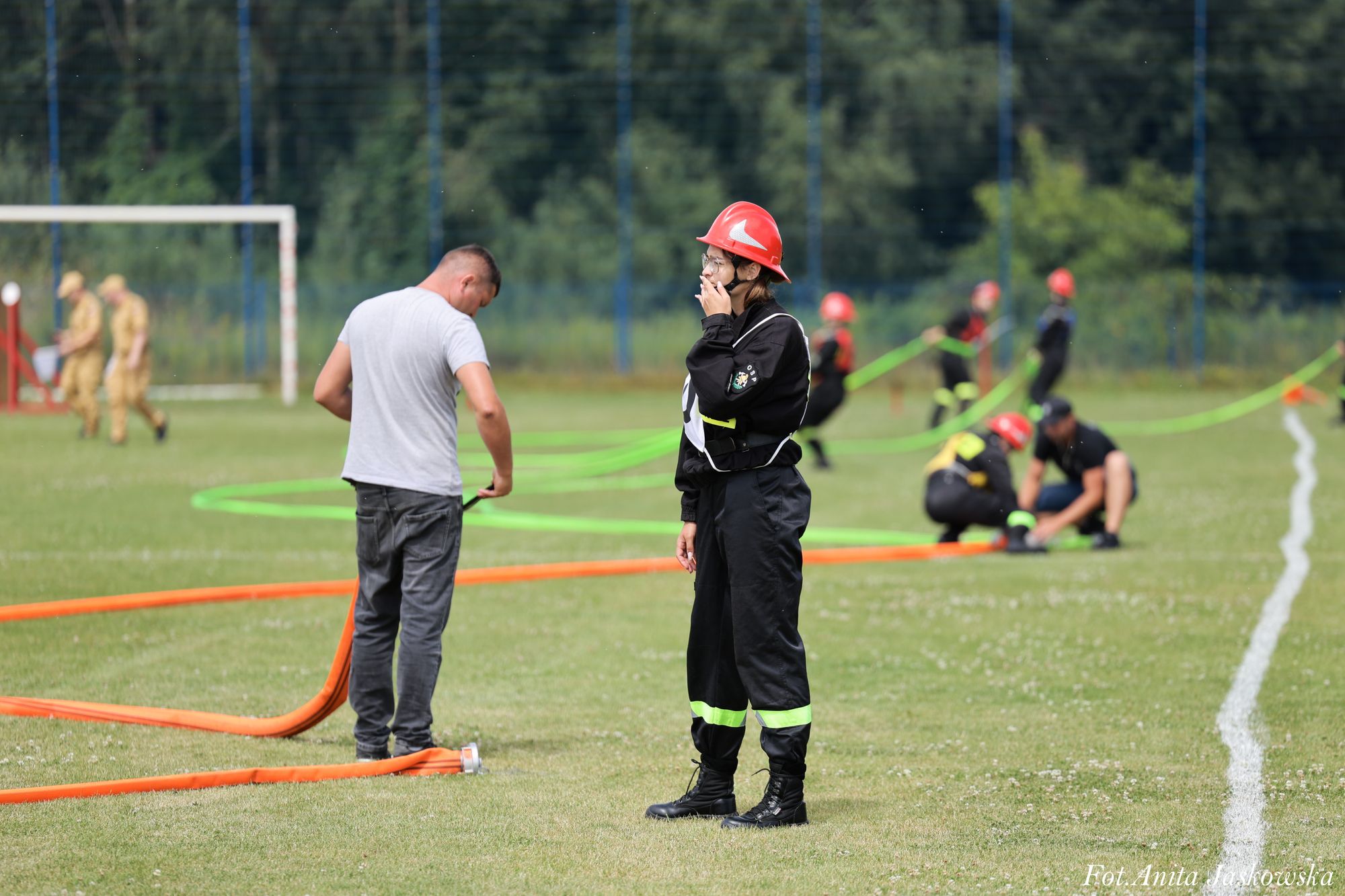 Kobieta w czarnym stroju ochronnym i czerwonym kasku pali papierosa na trawiastym boisku, mężczyzna obok trzyma pomarańczową rurę, w tle ludzie z czerwoną ochroną i zielonymi wężami.
