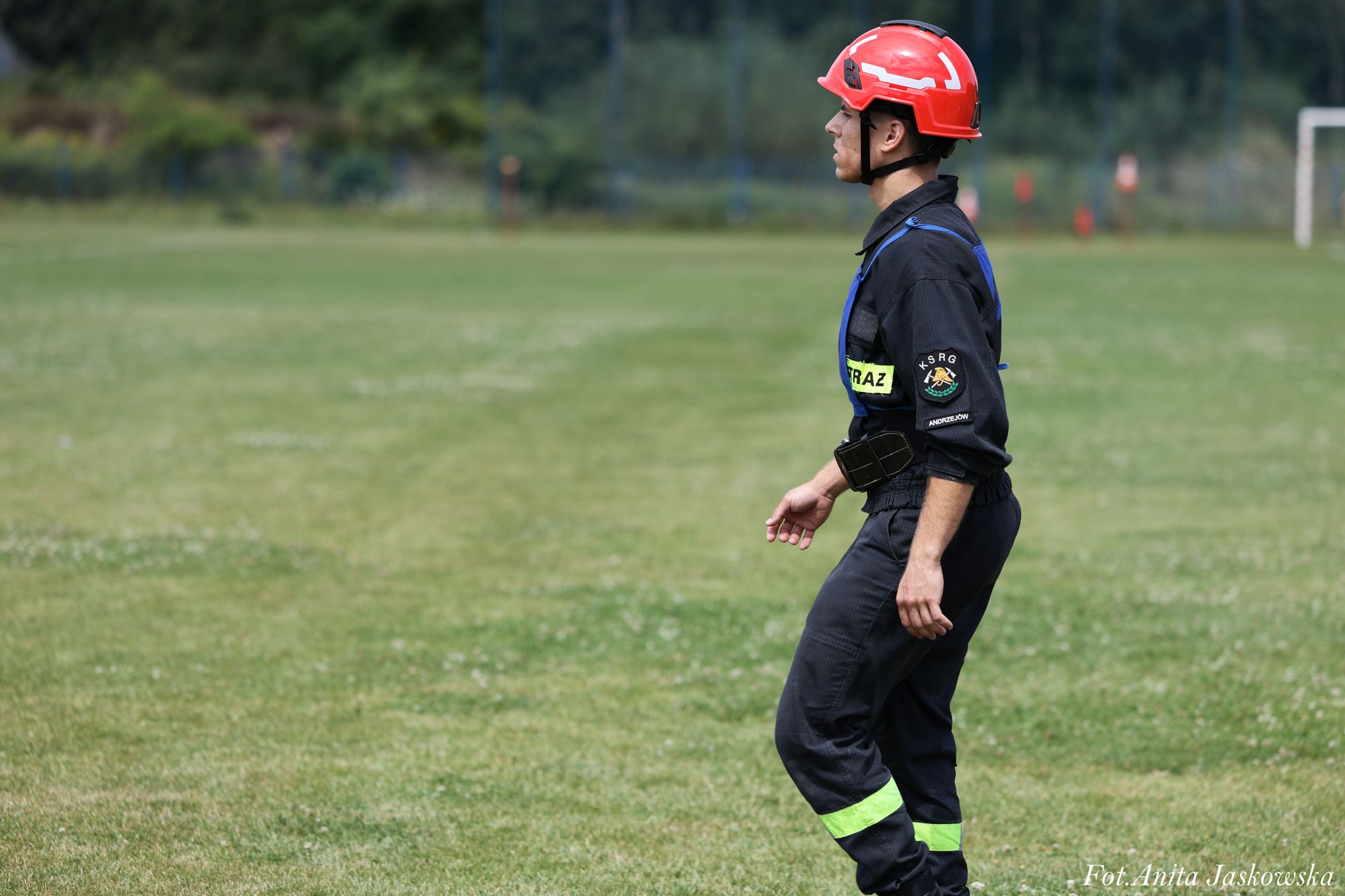 Mężczyzna w czarnym ubraniu strażaka z odblaskowymi paskami i czerwonym hełmem stoi na zielonym trawiastym boisku.