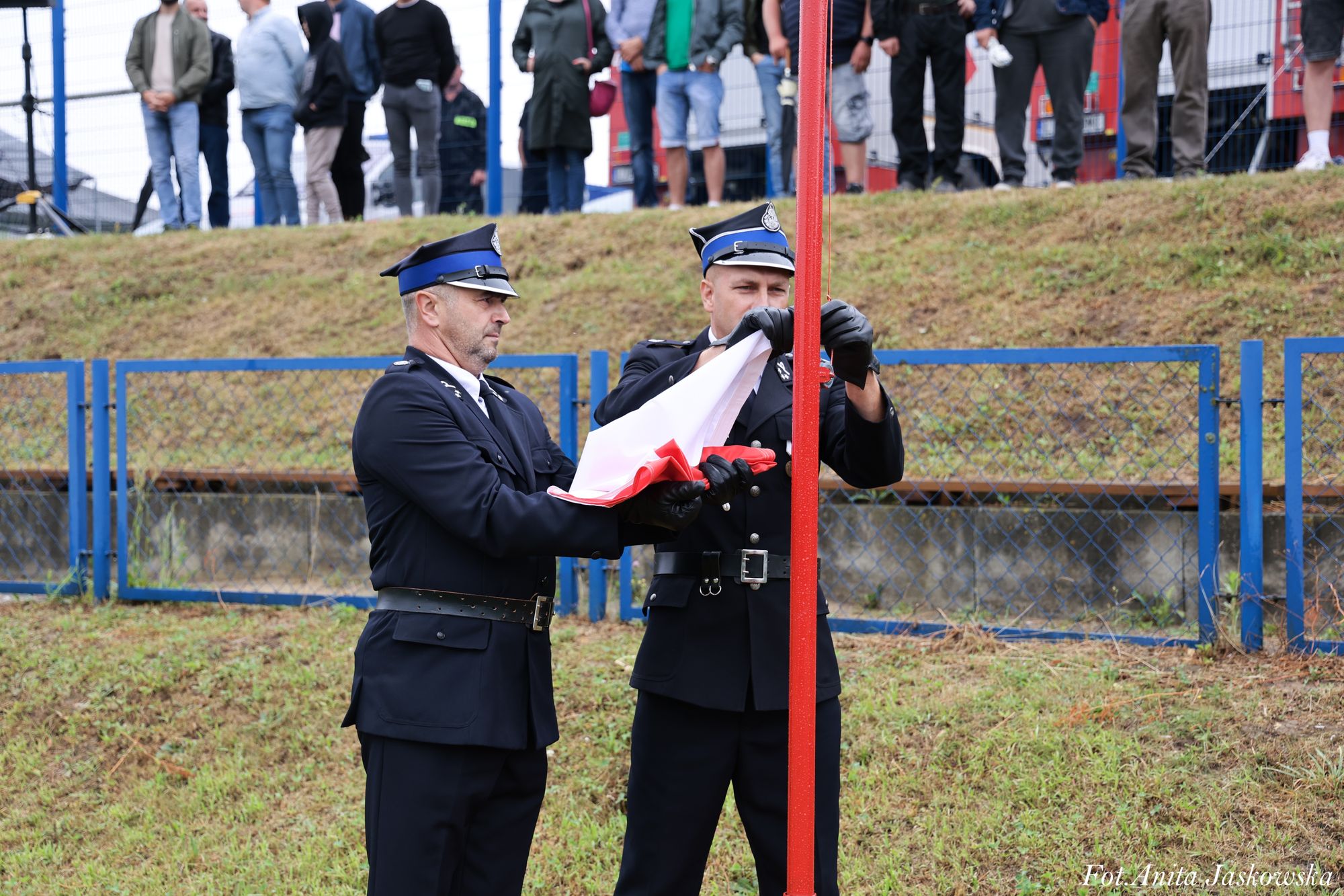 Dwóch mężczyzn w granatowych mundurach z niebieskimi daszkami składa biało-czerwoną flagę przy czerwonym maszcie, w tle zielone zbocze i niebieski płotek.