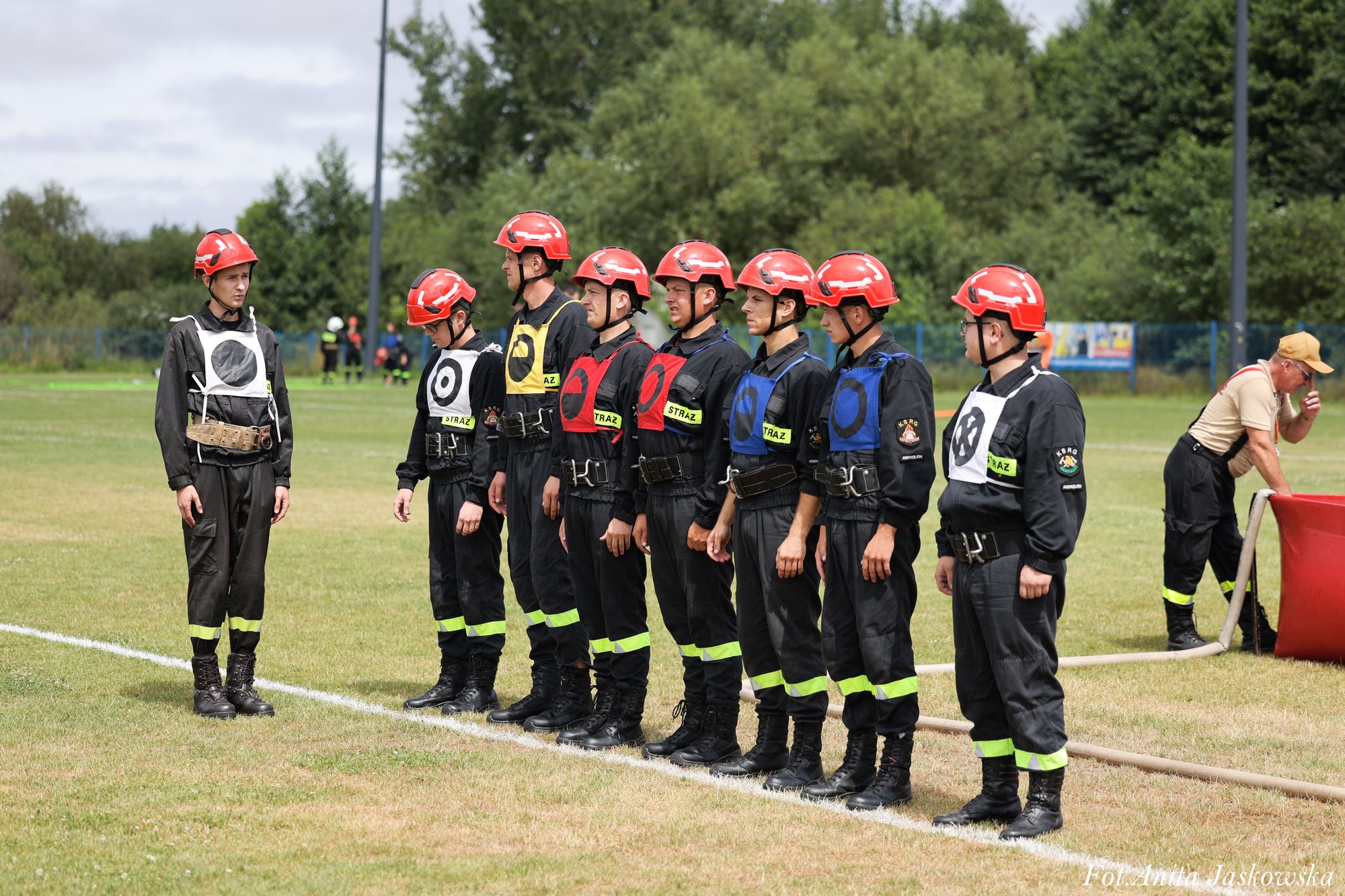 Osiem osób w czarnych mundurach z odblaskowymi paskami i czerwonych kaskach, stojących w szeregu na trawniku, jedna osoba po lewej w podobnym stroju.